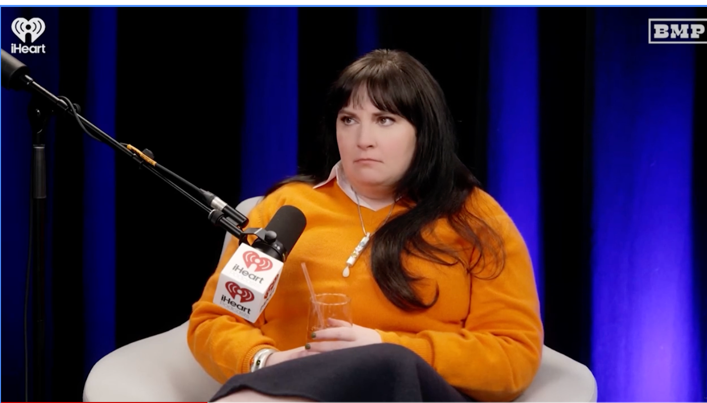 Person sitting connected  a seat  with a microphone, wearing a long-sleeve top, holding a drink, looks pensive. iHeart and BMP logos visible