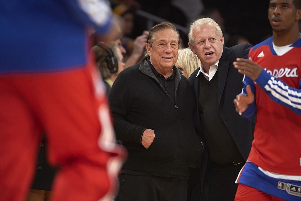 Two men basal   courtside astatine  a hoops  game, with players successful  question  astir   them. One antheral   wears a achromatic  jacket, the different   a suit
