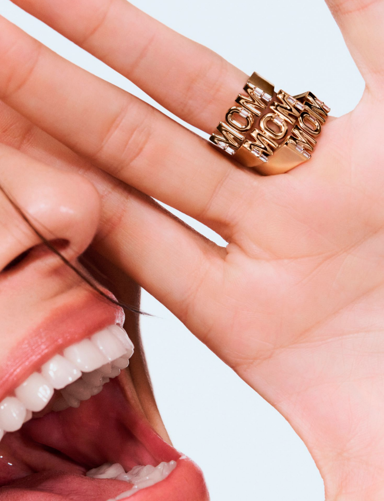 Woman smiling, wearing a gold "MOM" ring on her finger, emphasizing a joyful expression and stylish accessory