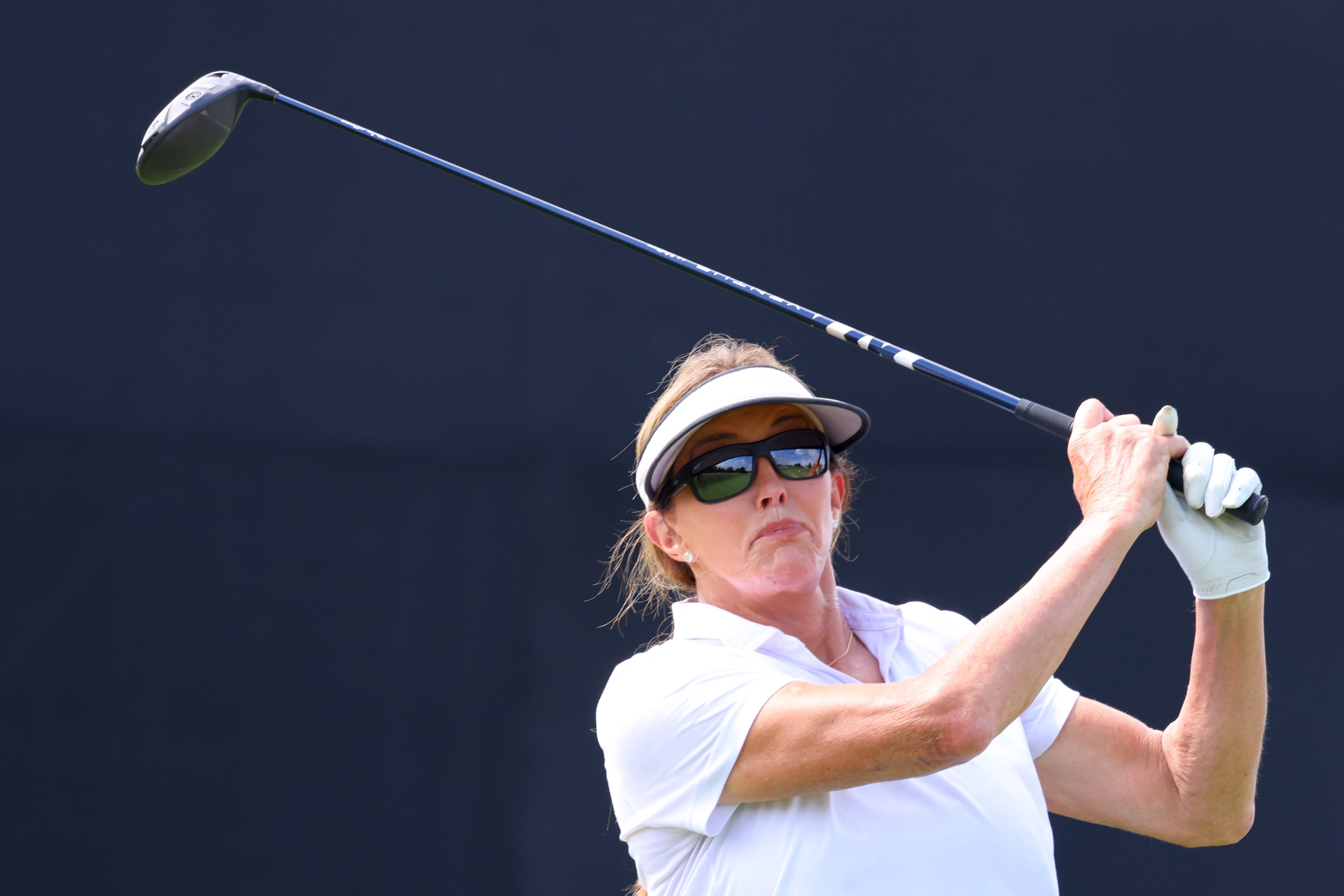 Caitlyn Jenner playing golf, wearing a visor and sunglasses, holding a play  nine  mid-swing connected  a sunny day