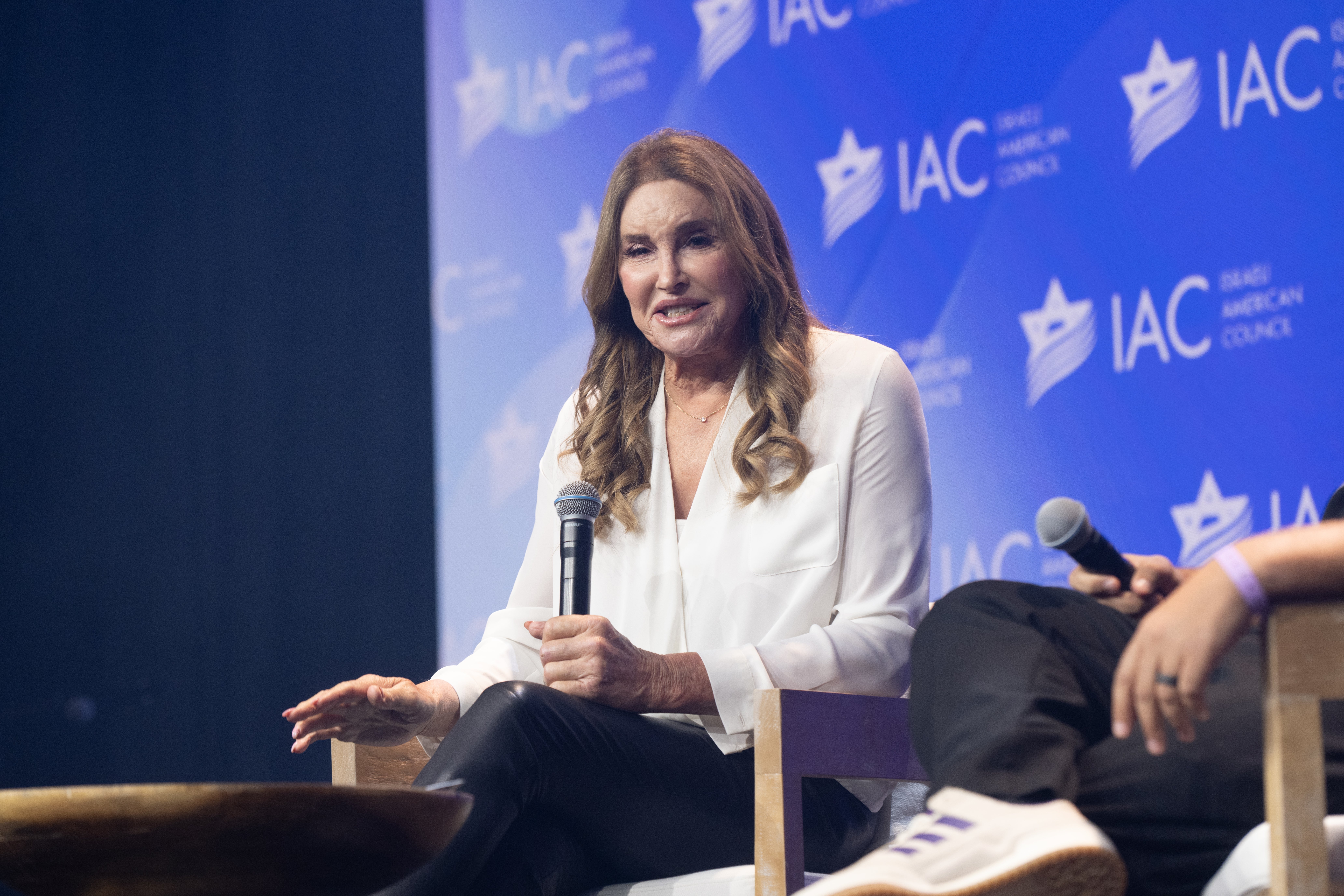Caitlyn Jenner speaking connected  signifier    astatine  a sheet  event, wearing a blouse and pants, holding a microphone with IAC branding successful  the background