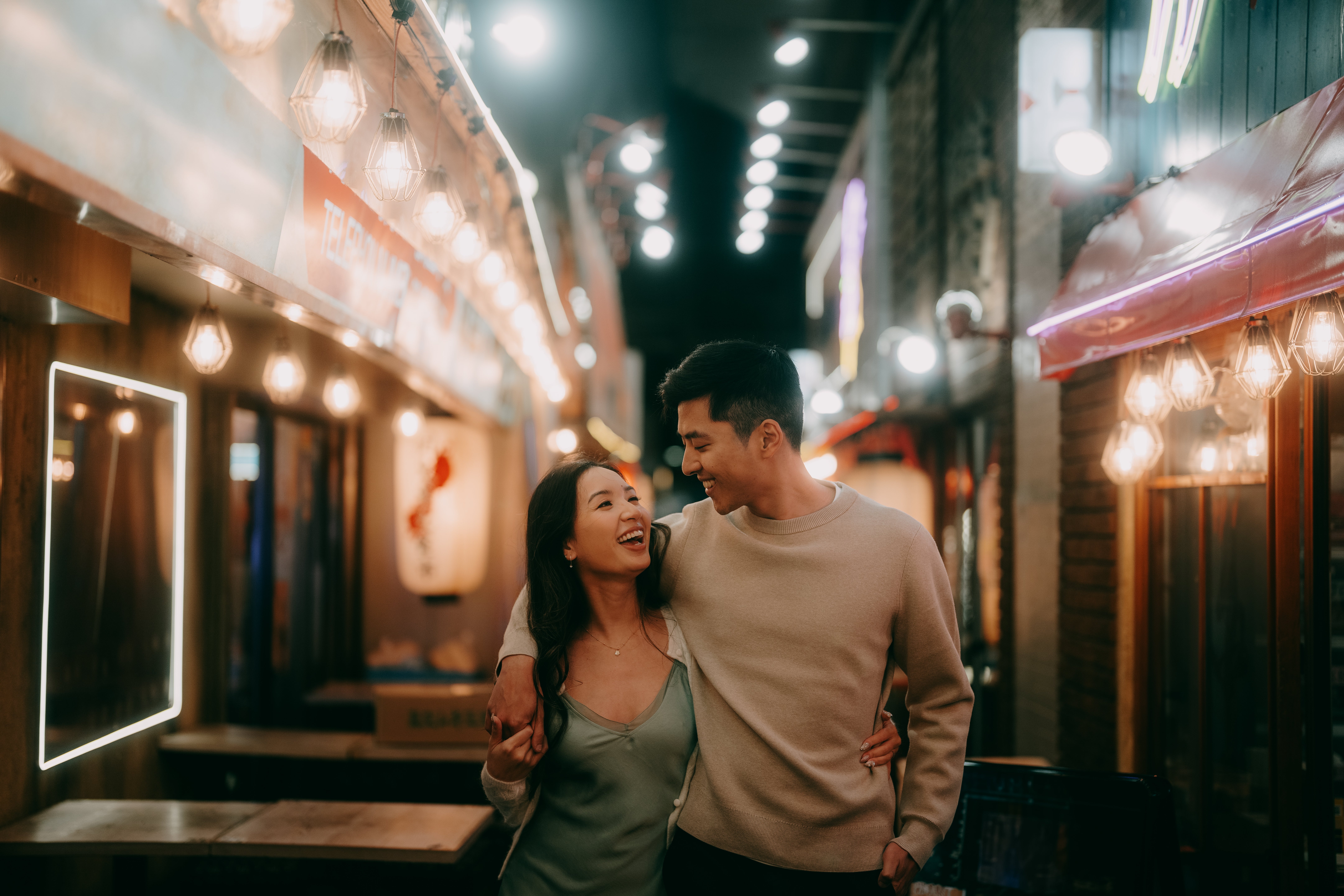 A couple walking and smiling at each other in a warmly lit alley at night, surrounded by string lights and neon signs