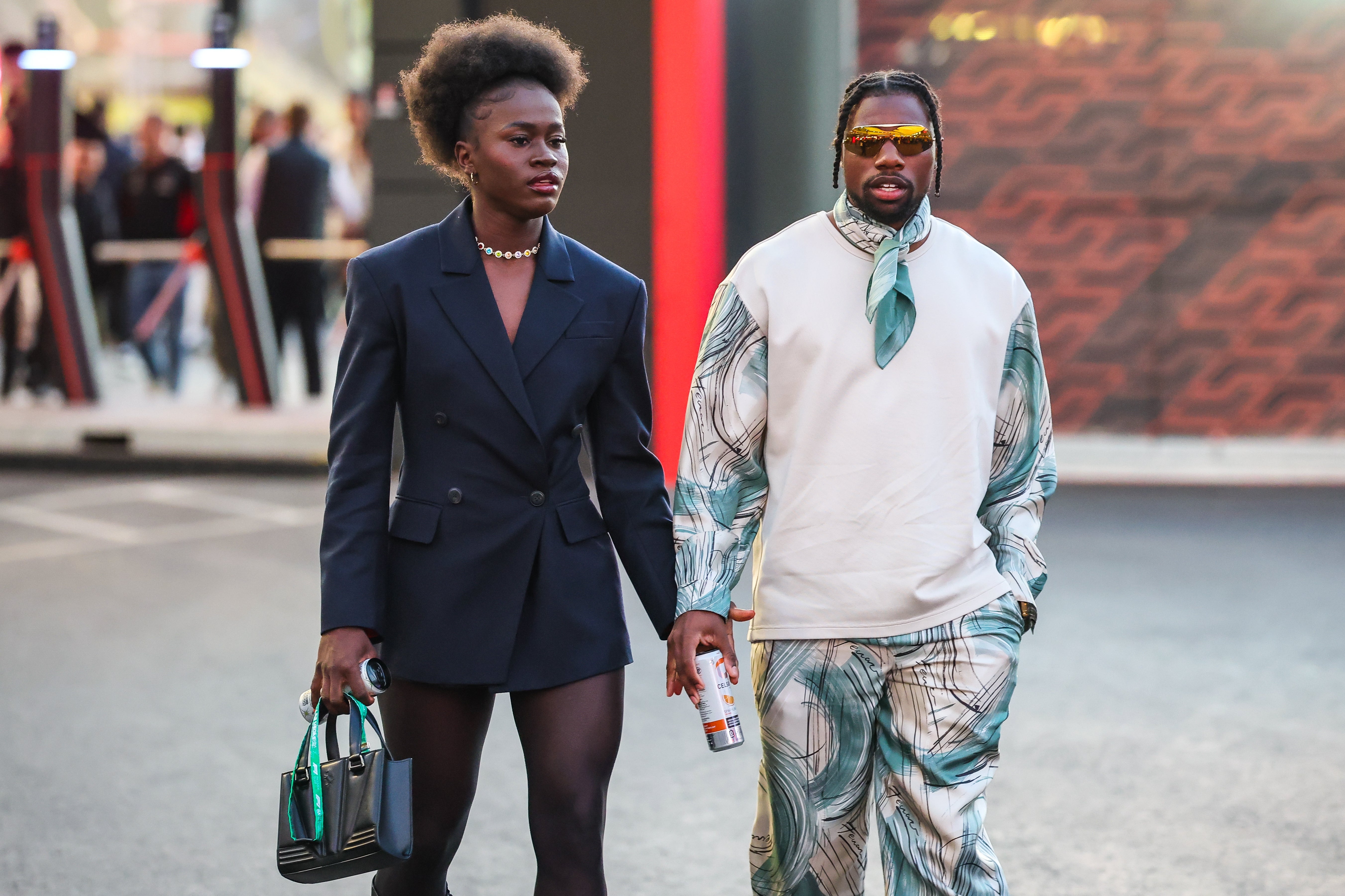 Two individuals walking. One wears a tailored blazer with shorts and pearl necklace, the different   successful  a patterned outfit with sunglasses