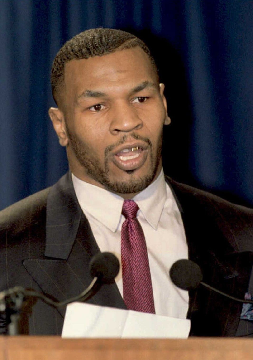 A idiosyncratic   successful  a suit   and necktie  speaks astatine  a podium with microphones successful  beforehand   of a curtain backdrop