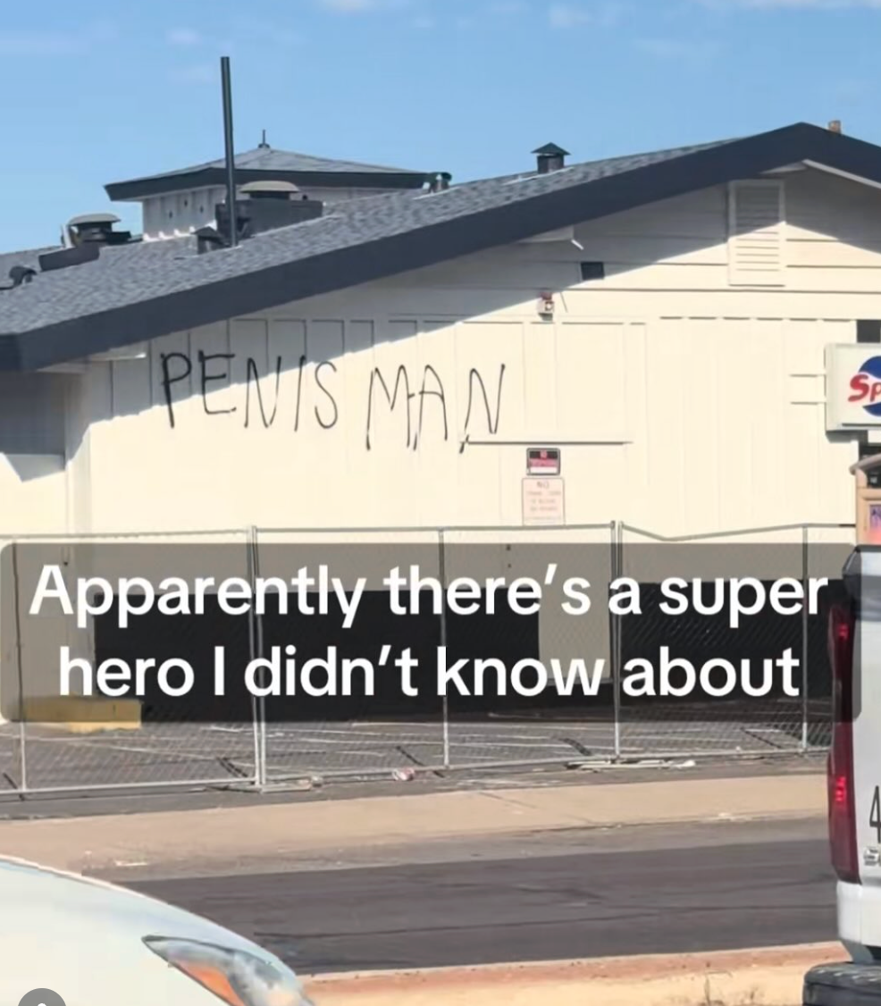 Graffiti on a building reads "Penis Man." Overlay text humorously says, "Apparently there's a superhero I didn't know about."