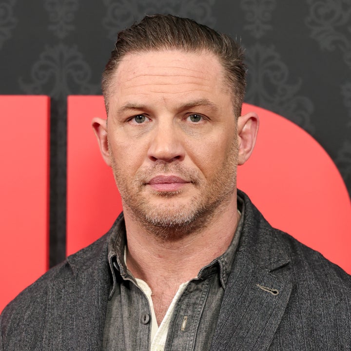 A person stands in front of a patterned backdrop, wearing a dark denim shirt and a pinstriped jacket