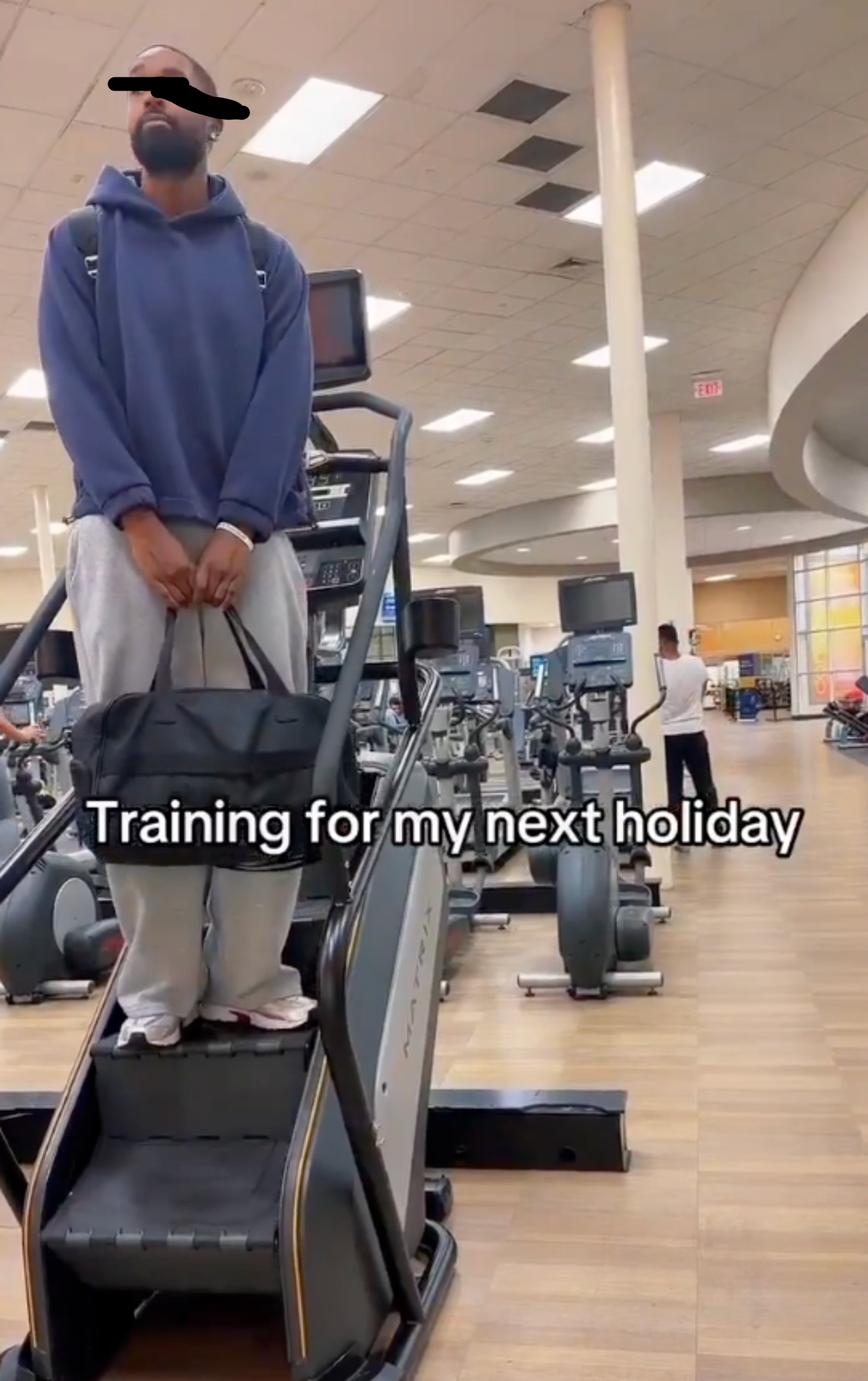 Person standing on a stair machine in a gym, wearing casual workout clothes, holding a bag. Text overlay says: "Training for my next holiday."