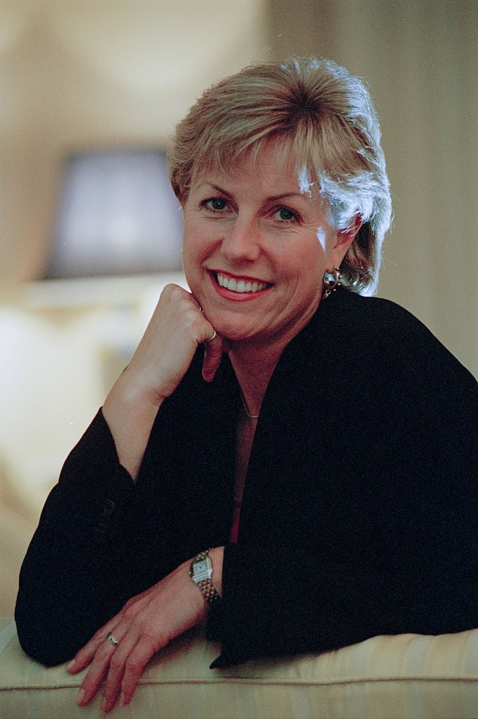 Person smiling, seated, resting chin connected  hand, wearing a achromatic  overgarment   and watch, with a relaxed and approachable expression