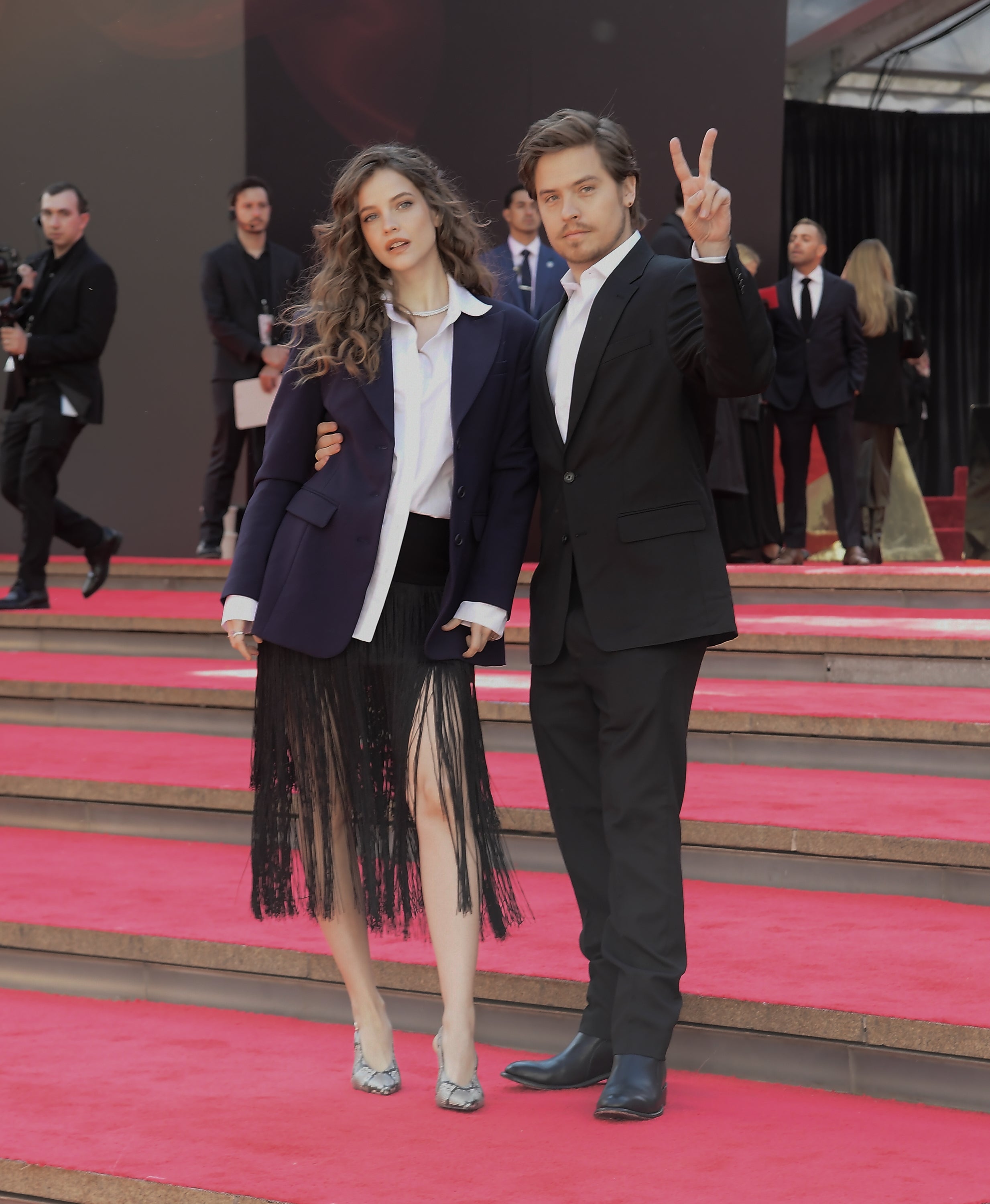 A antheral   successful  a suit   makes a bid    motion   beside a pistillate   with curly hair, wearing a blazer and fringe skirt, connected  a reddish  carpet