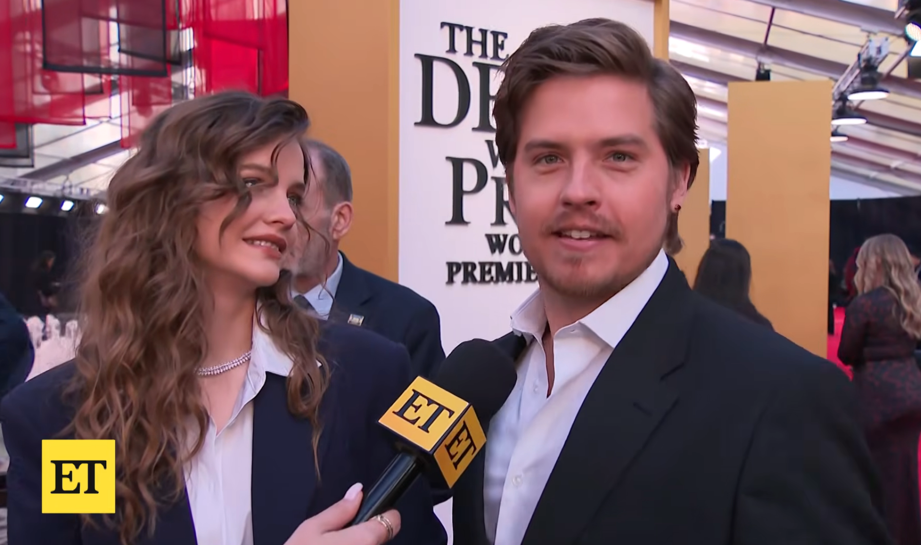 A antheral   and pistillate   are interviewed connected  the reddish  carpet. The antheral   wears a suit, and the pistillate   has wavy hairsbreadth  and a chic outfit