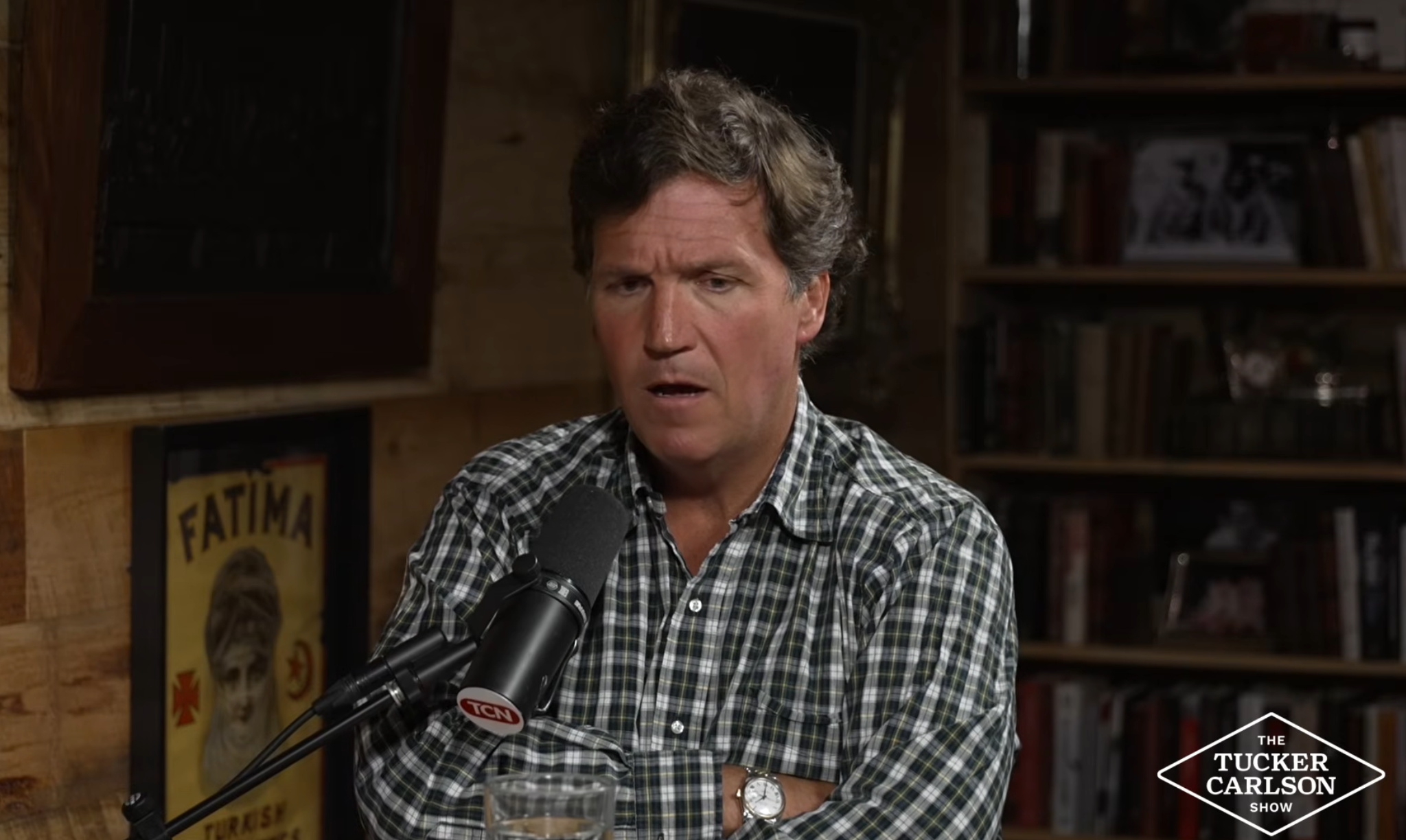 Person successful  plaid garment  speaking into a microphone connected  "The Tucker Carlson Show" set. Books and framed pictures successful  the background