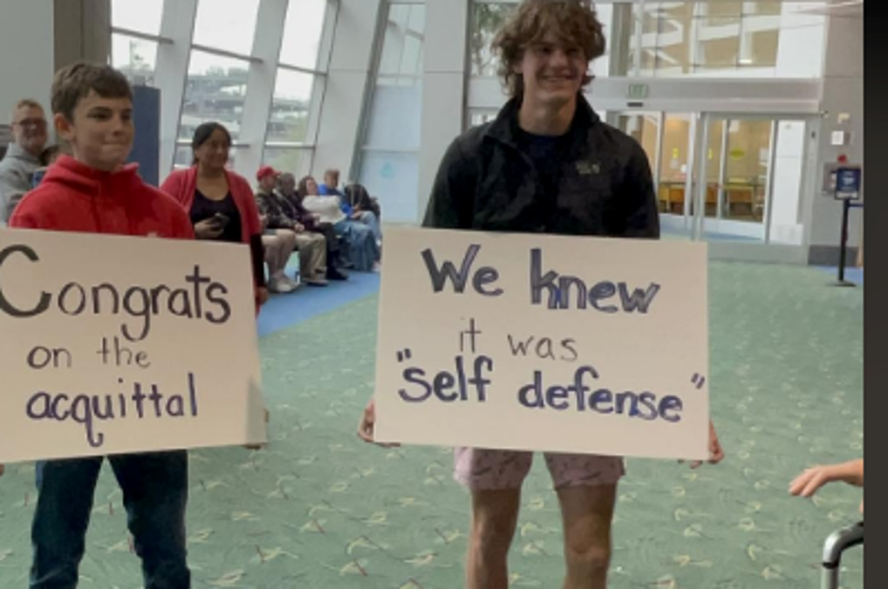 Two people in an airport hold signs: "Congrats on the acquittal" and "We knew it was 'self defense.'"
