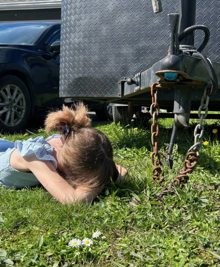Child resting on grass next to a trailer hitch, with a car in the background and daisies nearby