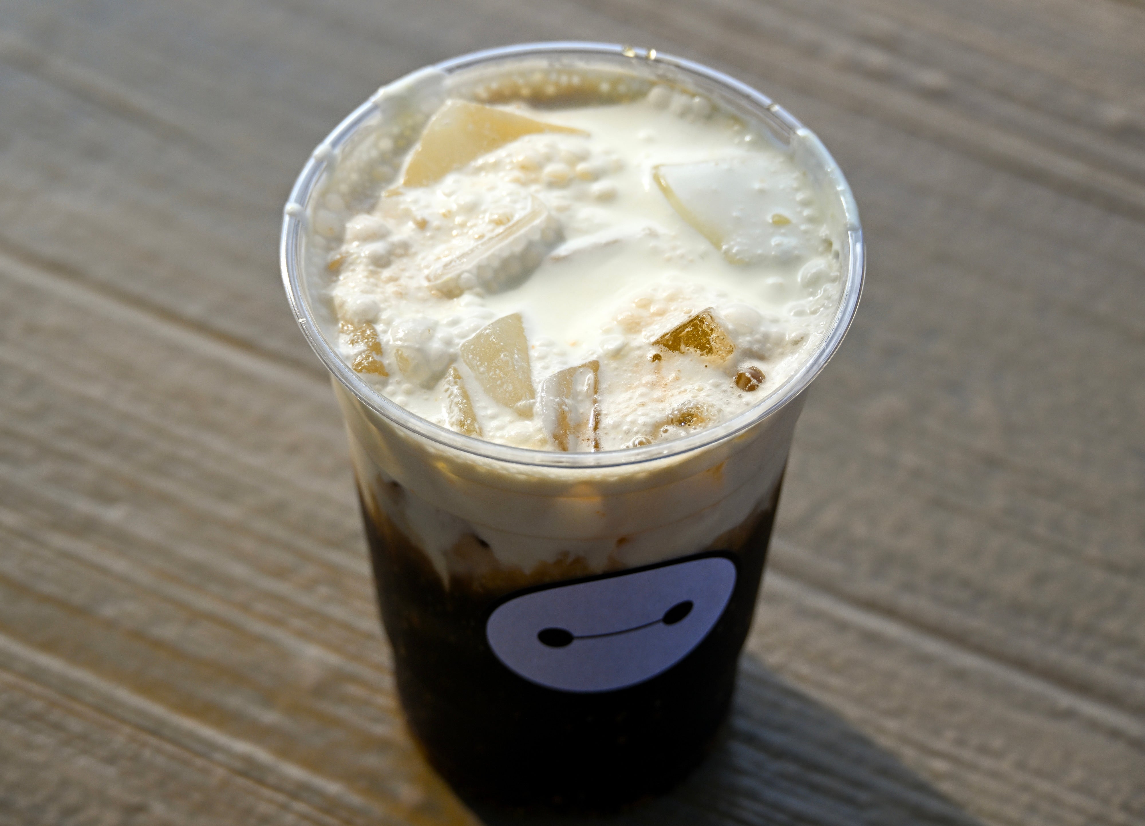 Cup of iced java  with cream, topped with taro chunks, placed connected  a textured woody  surface. Cup has a playful elemental  look   graphic