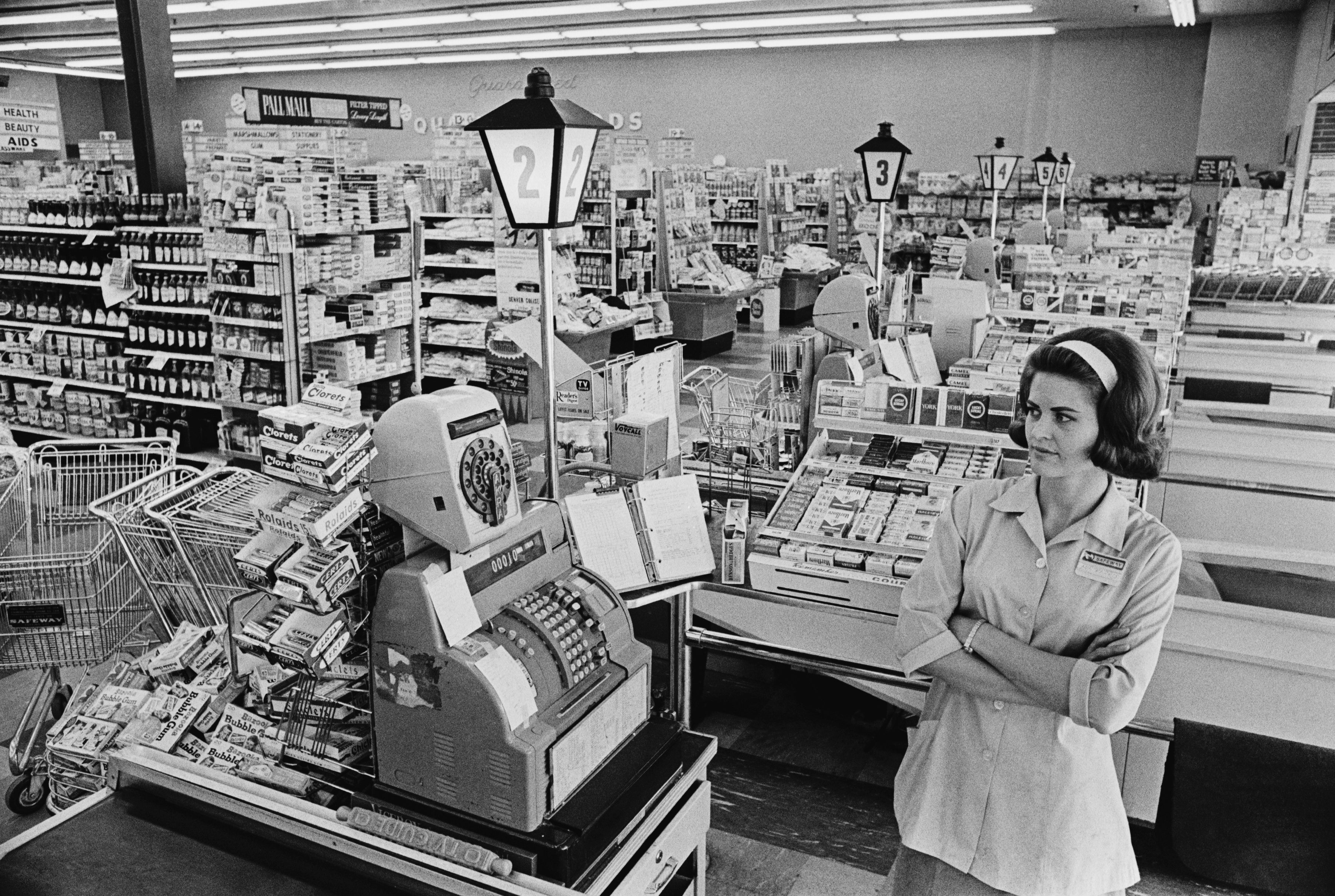 Vintage photograph  of a 1960s supermarket checkout country  with a cashier lasting  beside a mechanical registry  amid assorted  groceries and products