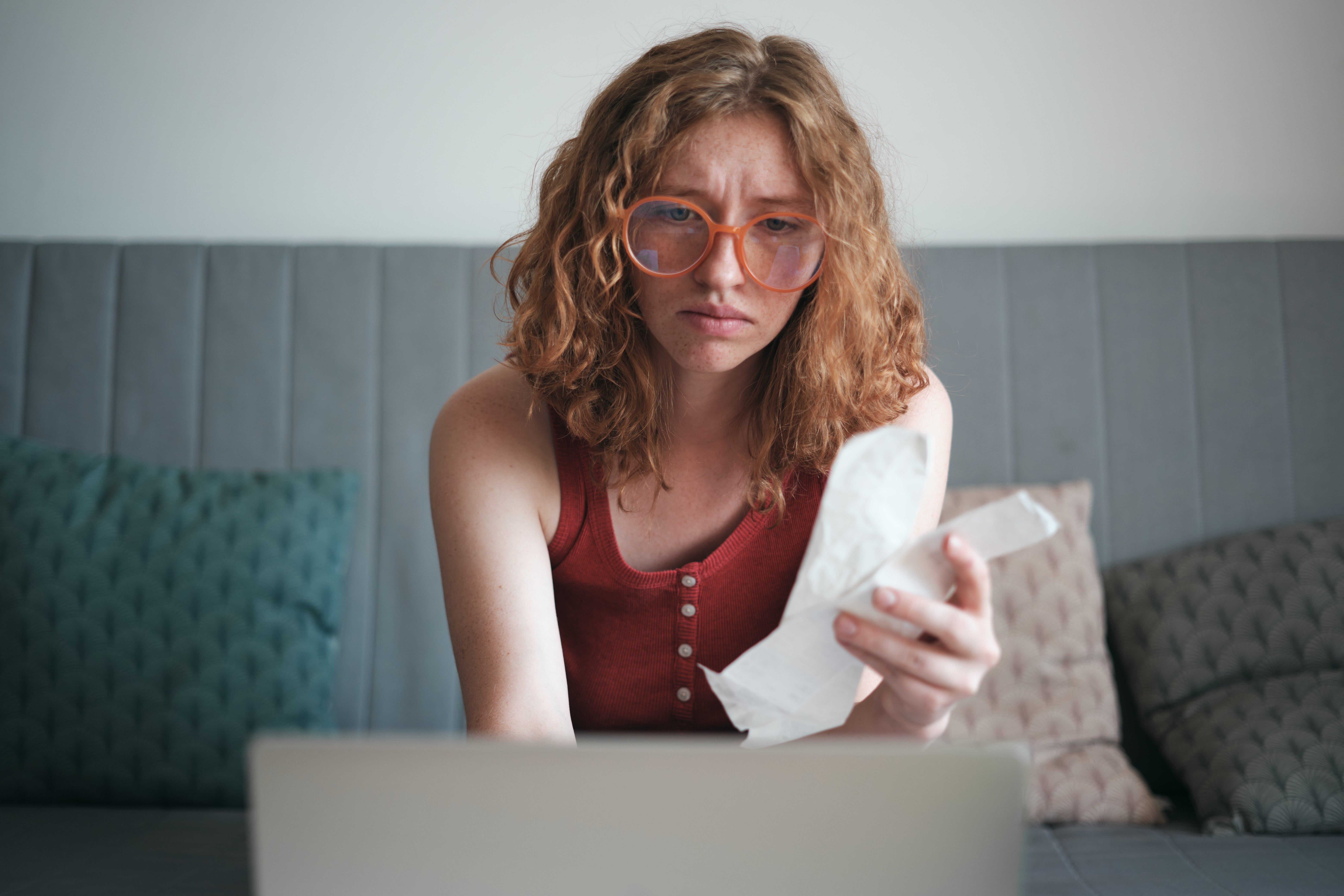 Person with curly hairsbreadth  and glasses looking acrophobic   astatine  a receipt portion    sitting successful  beforehand   of a laptop connected  a grey  sofa