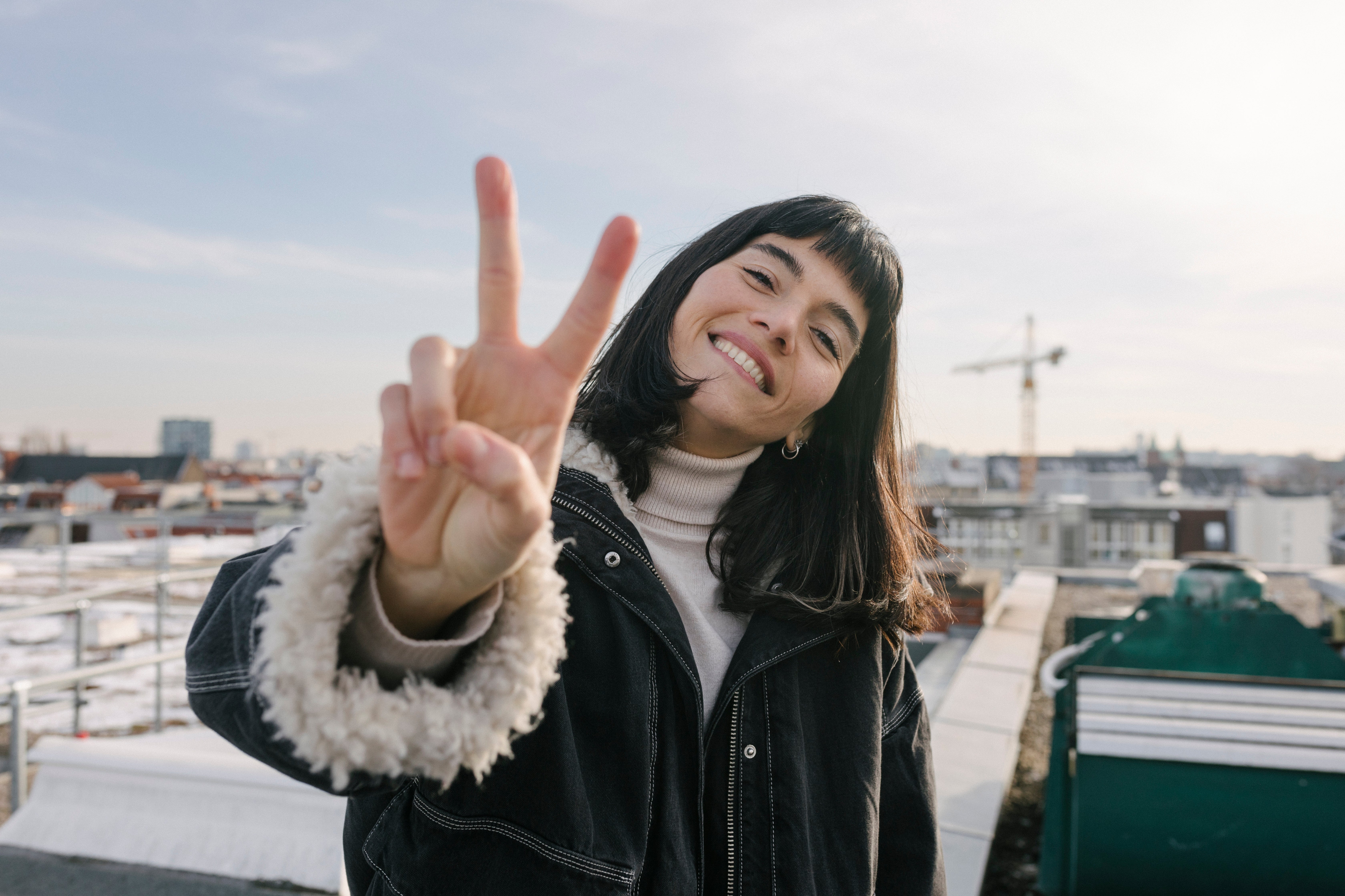Person smiling connected  a rooftop, wearing a overgarment   with furry cuffs, making a bid    sign. Cityscape and operation  crane successful  the background