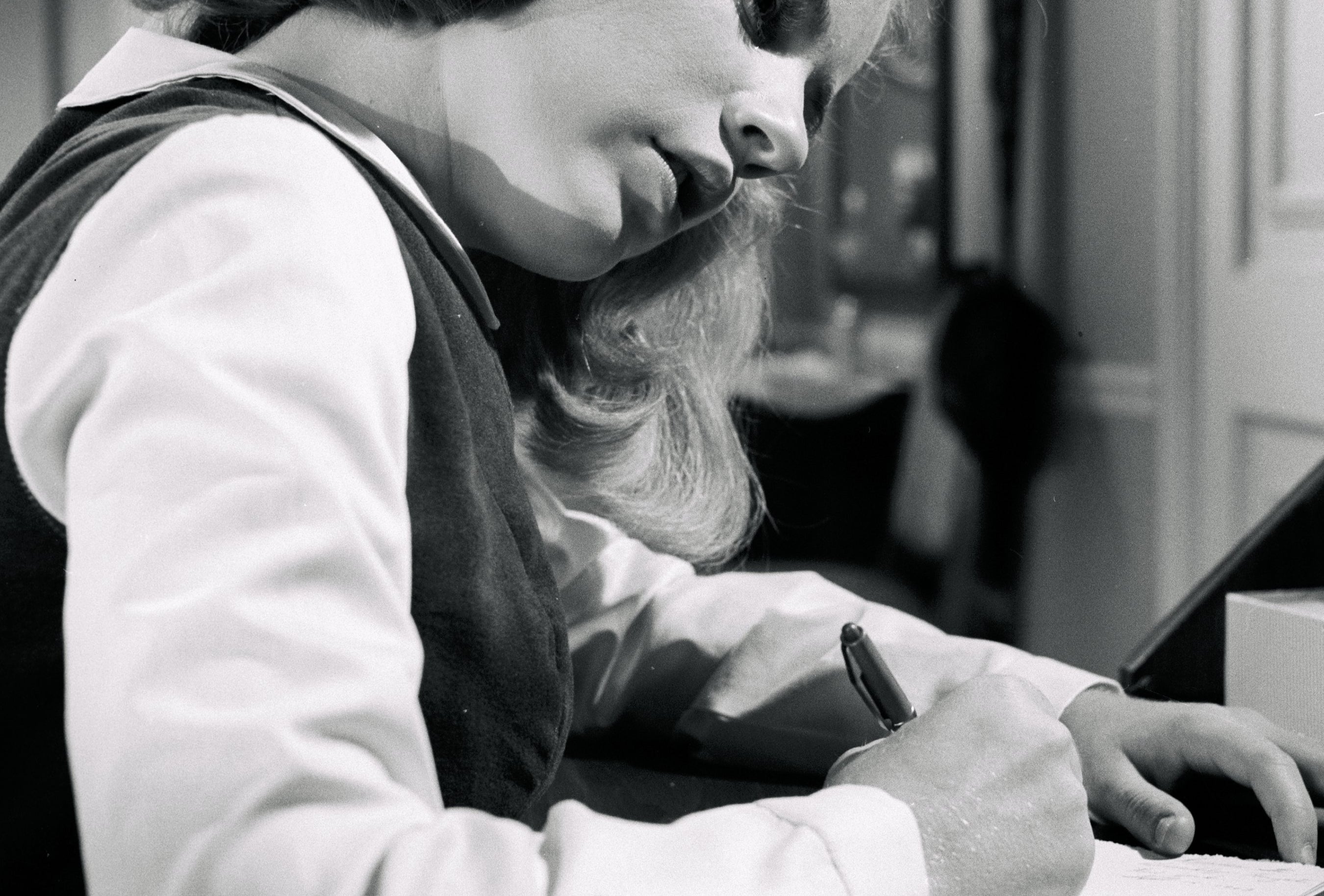 A young pistillate   successful  vintage attire writes intently astatine  a desk, evoking a mid-20th-century setting