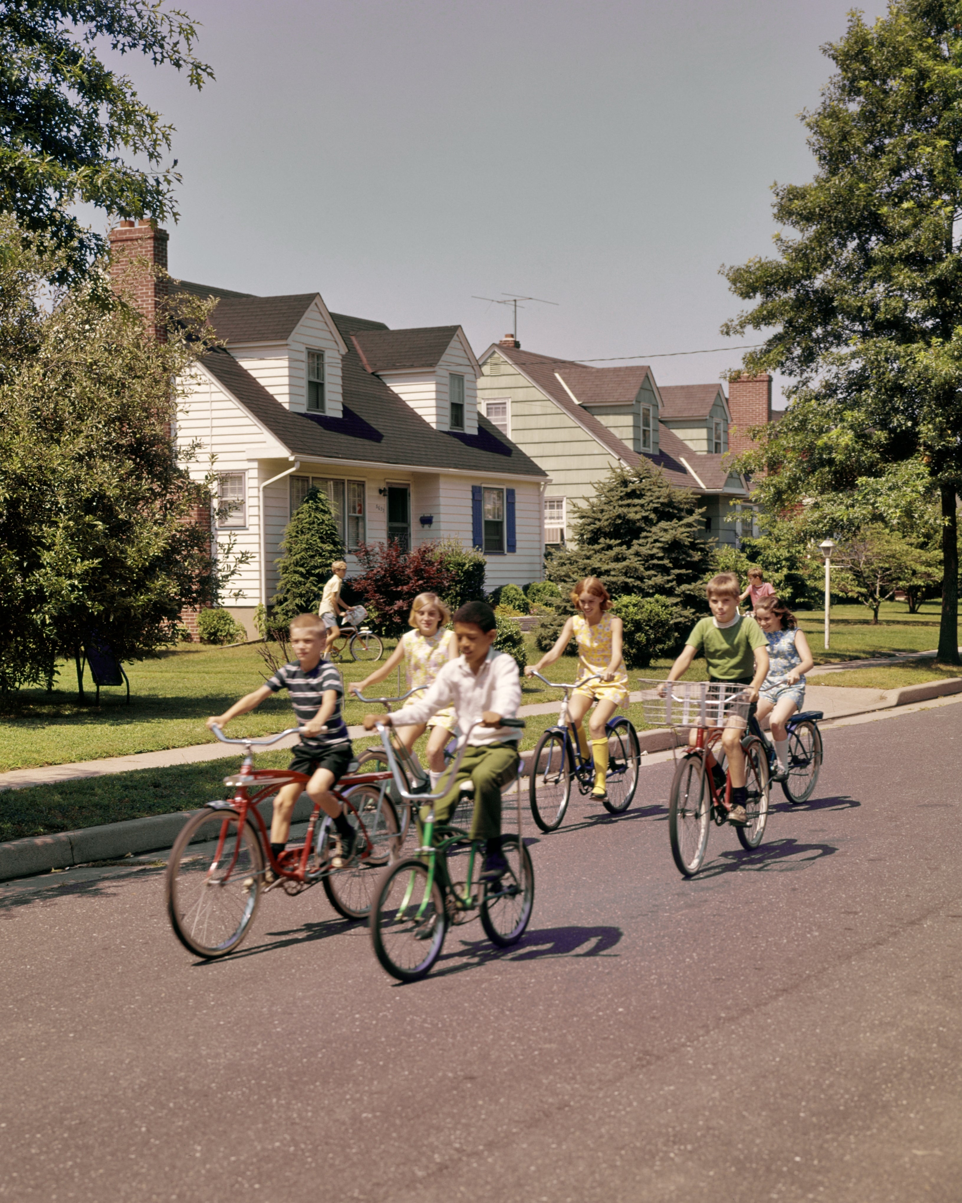 A radical  of children joyfully thrust   bicycles down   a suburban thoroughfare  lined with houses, trees, and a wide   sky, capturing a retro, carefree scene