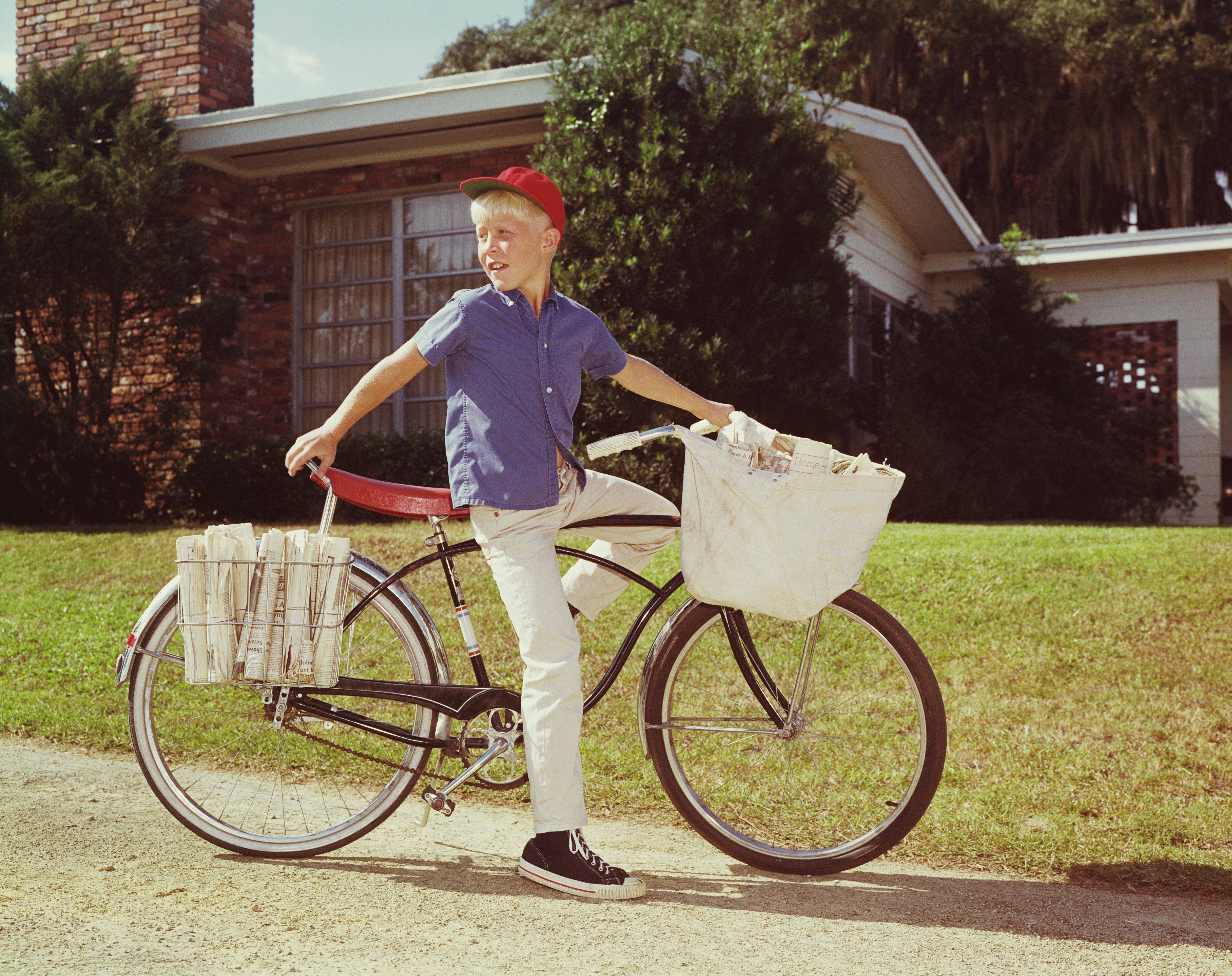 A young lad  successful  a headdress  is connected  a bicycle with newspapers successful  the basket, acceptable   for transportation  successful  a suburban neighborhood