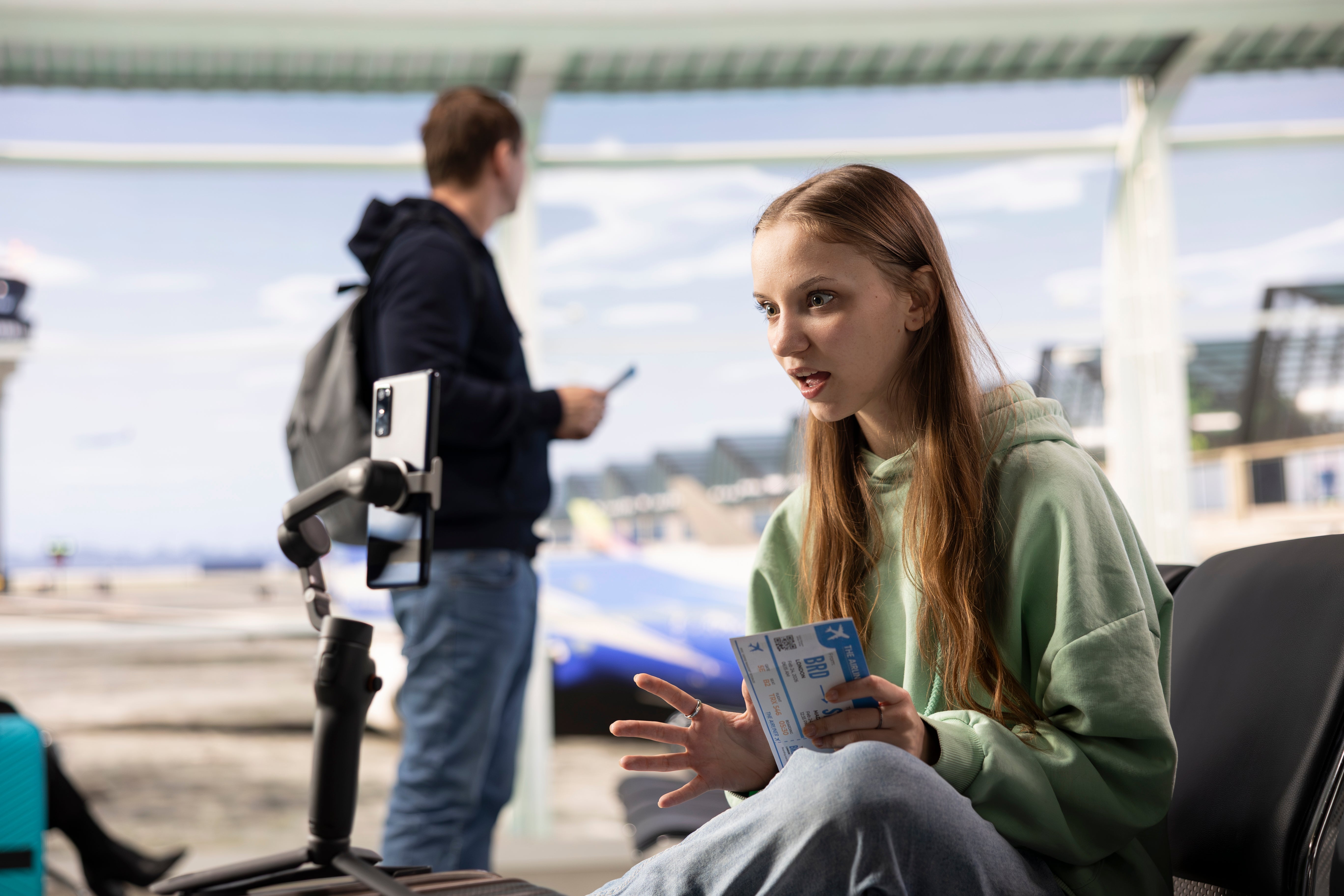 A pistillate   successful  casual covering  talks to her telephone  camera astatine  an airport, holding tickets; a antheral   with a backpack is successful  the background