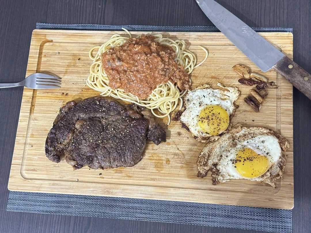 Steak, spageti dengan saus tomat, dan telur goreng disajikan di atas talenan kayu dengan pisau dan garpu di sampingnya