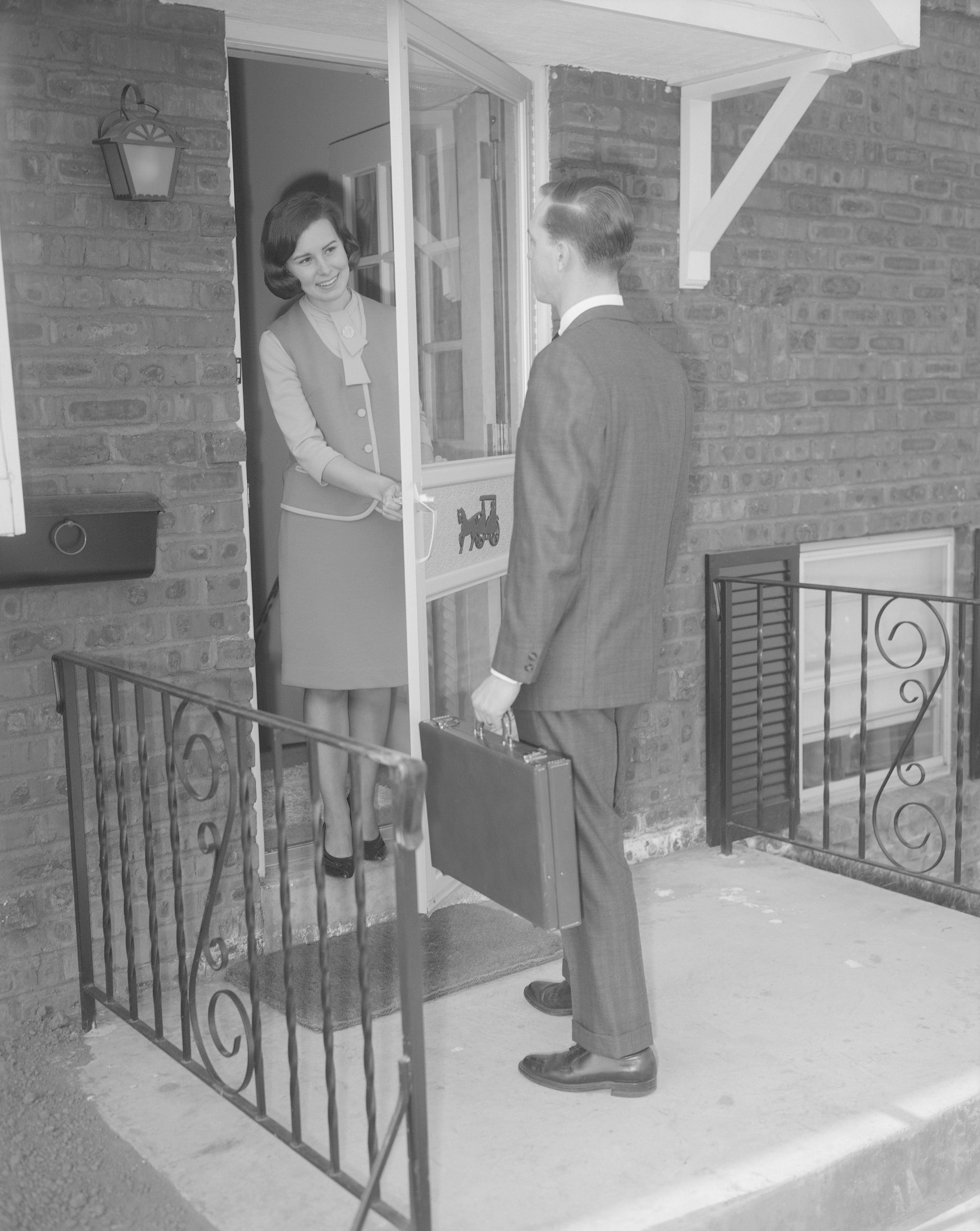  A pistillate   greets a antheral   successful  a suit   holding a briefcase astatine  the doorway  of a ceramic  home