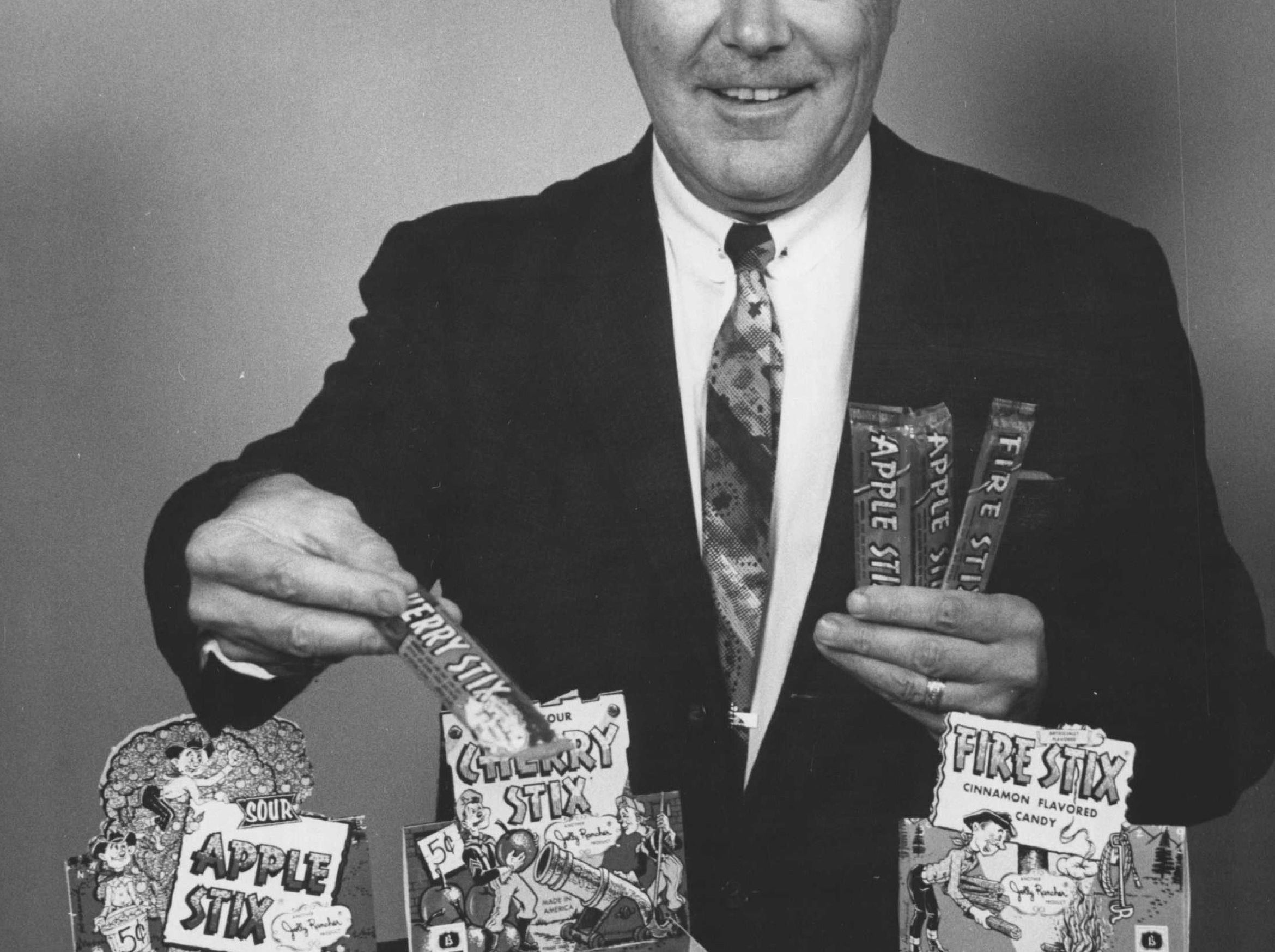 Man successful  a suit   presents vintage candy boxes labeled "Sour Apple Stix," "Cherry Stix," and "Fire Stix" with respective  sticks successful  hand