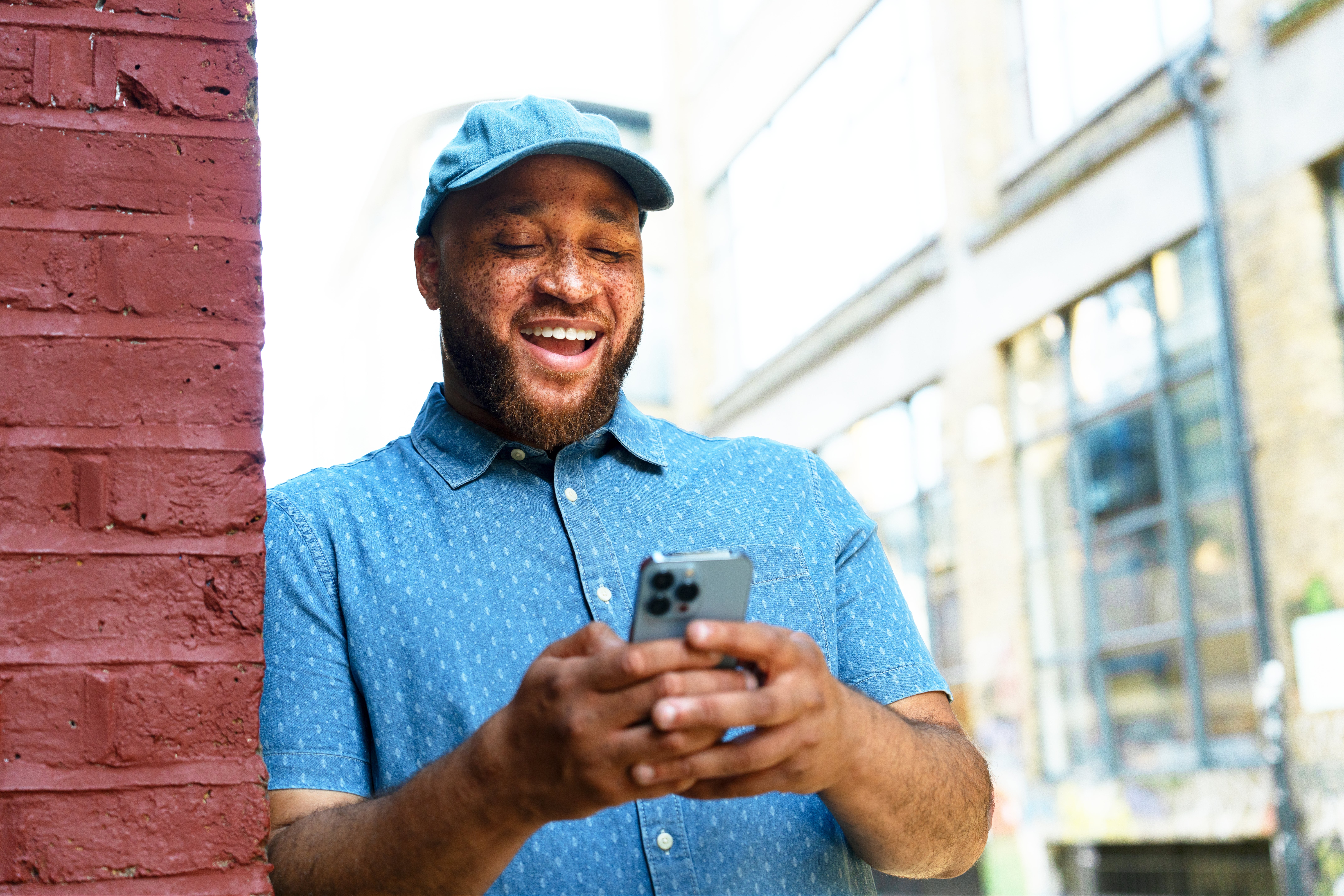 A idiosyncratic   successful  a casual garment  and headdress  leans against a ceramic  wall, smiling portion    looking astatine  a smartphone, outdoors successful  an municipality  setting