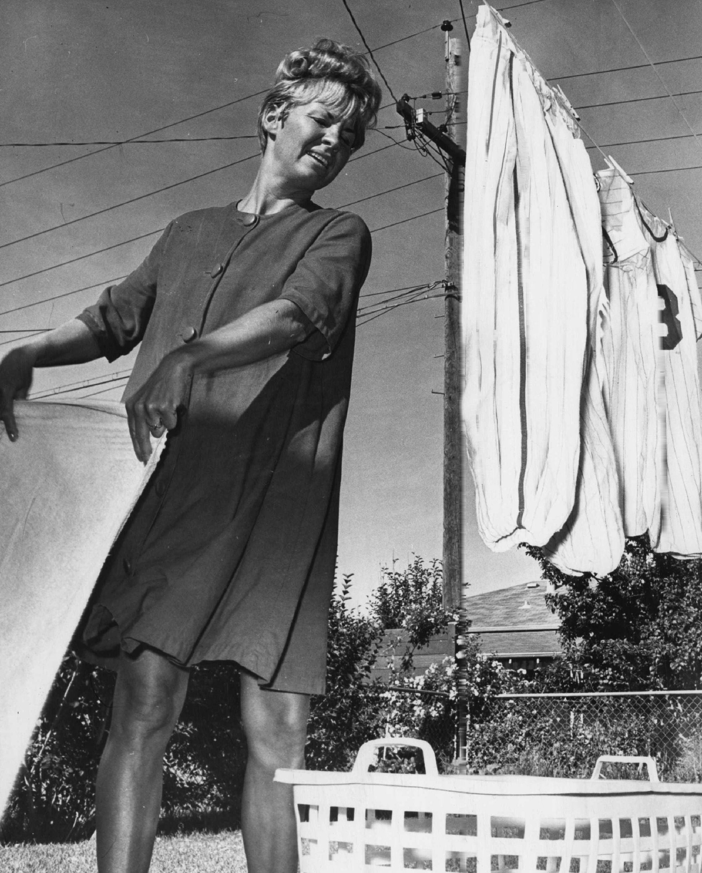 Woman smiling portion    hanging laundry outdoors adjacent  to a basket. Retro-style formal  suggests a classical  mid-20th period  setting