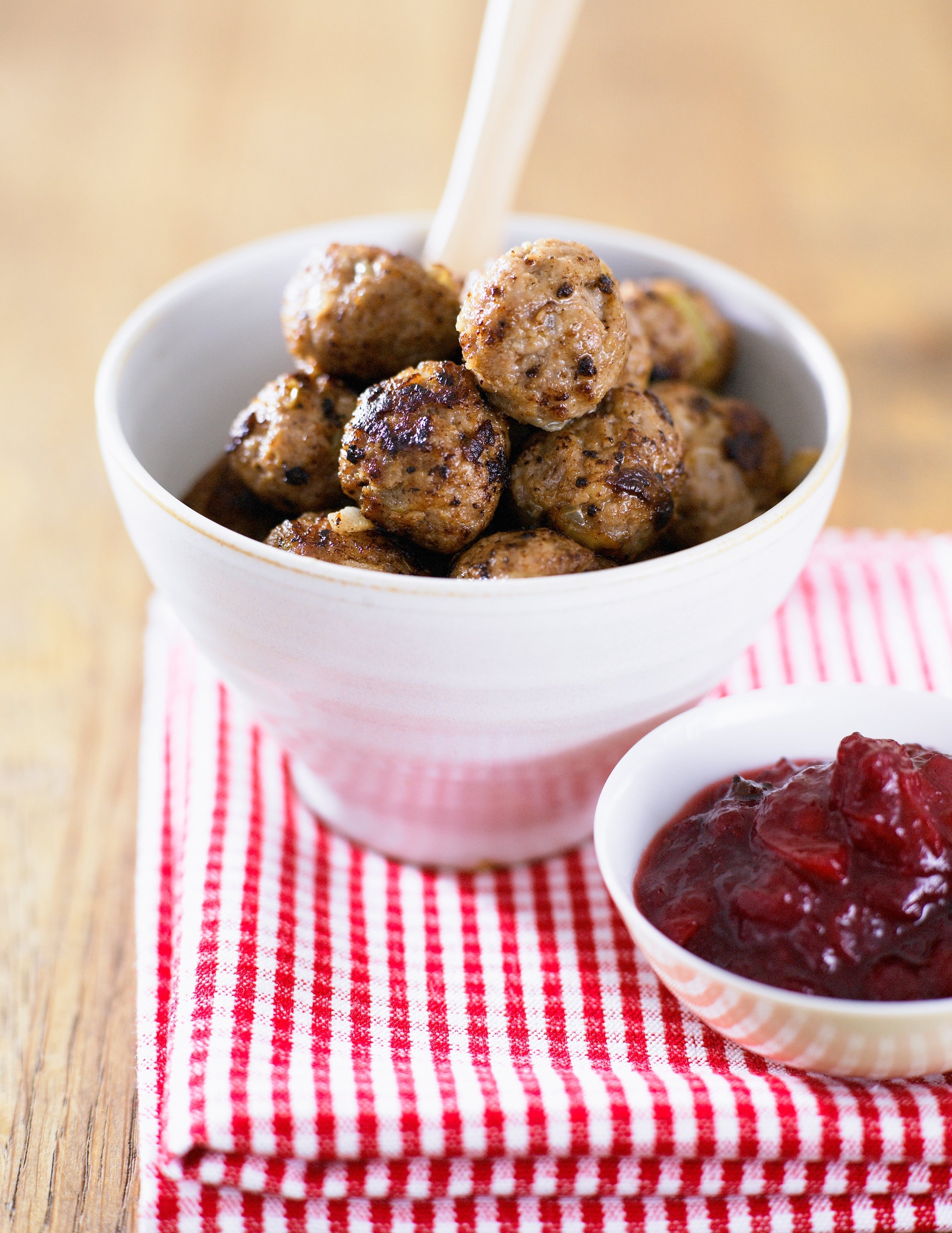 A vessel  of Swedish meatballs connected  a reddish  and achromatic  checkered cloth, accompanied by a tiny  crockery  of cranberry sauce