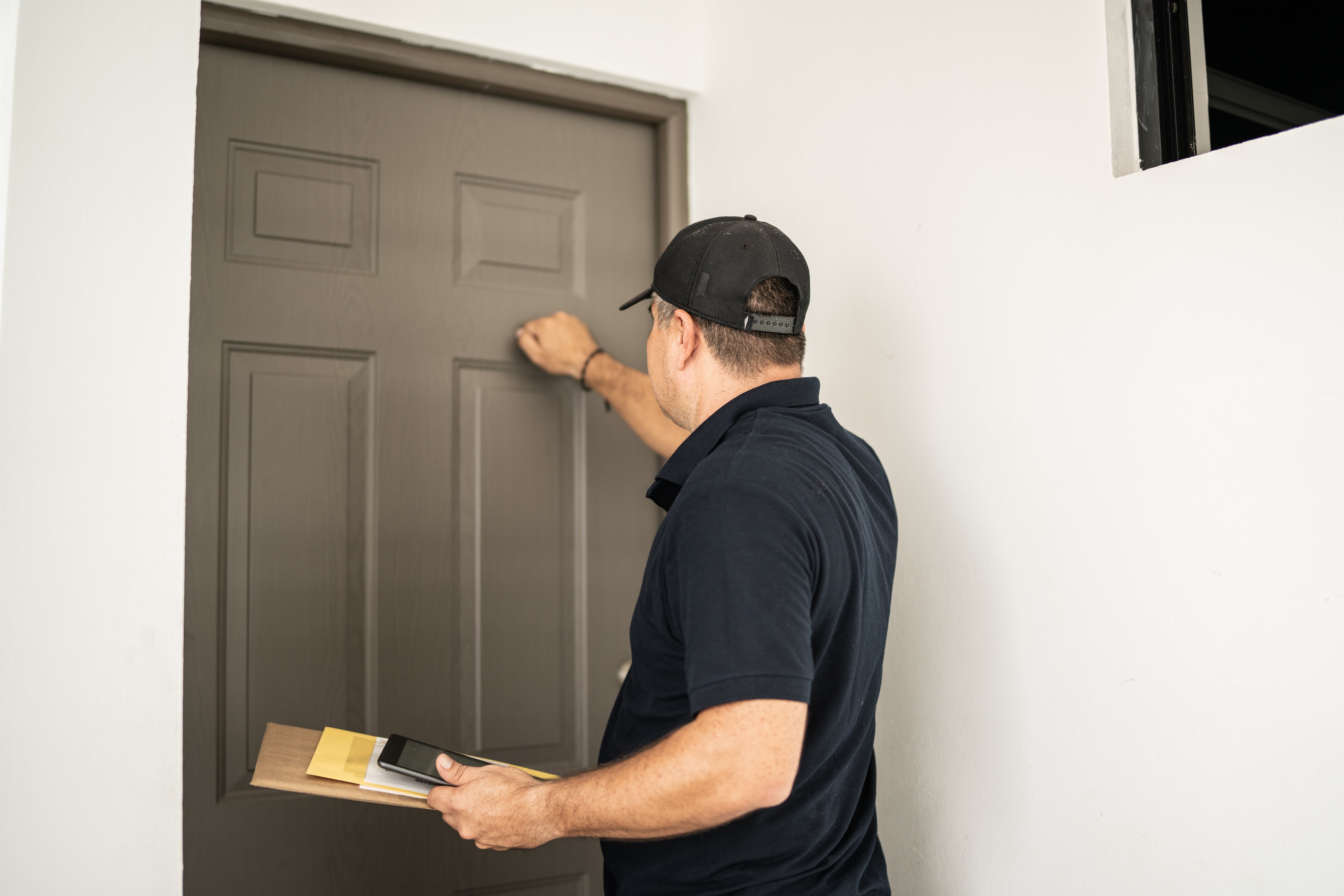 A transportation  idiosyncratic   successful  casual attire, holding a clipboard and envelope, knocks connected  a doorway  successful  a residential setting