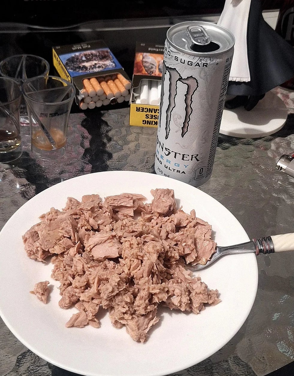 Sepiring tuna dengan garpu, dikelilingi minuman energi, rokok, dan gelas di atas meja
