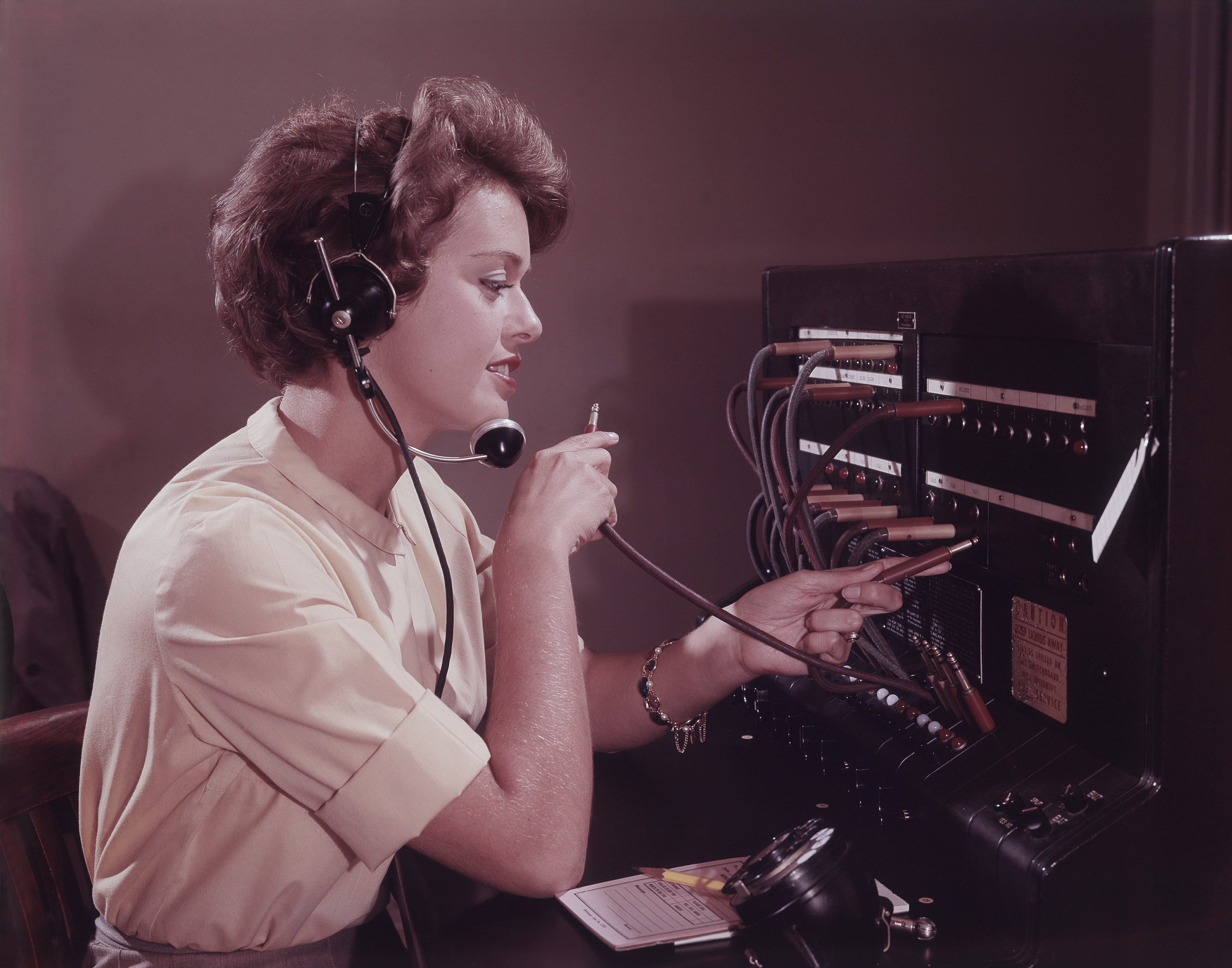 A pistillate   successful  vintage attire operates a 1960s-era telephone   switchboard, connecting calls manually