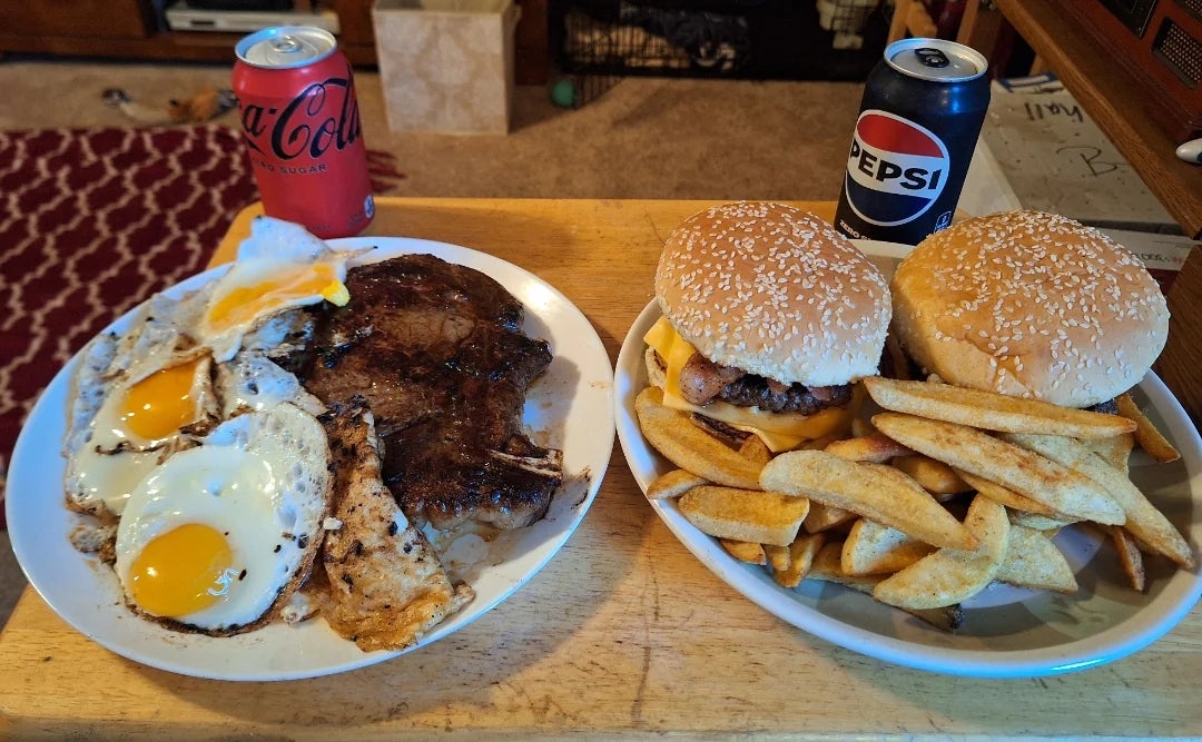 Dua kaleng soda dan dua piring makanan di atas meja kayu — satu berisi tiga telur goreng & steak dan satu dengan dua burger & kentang goreng