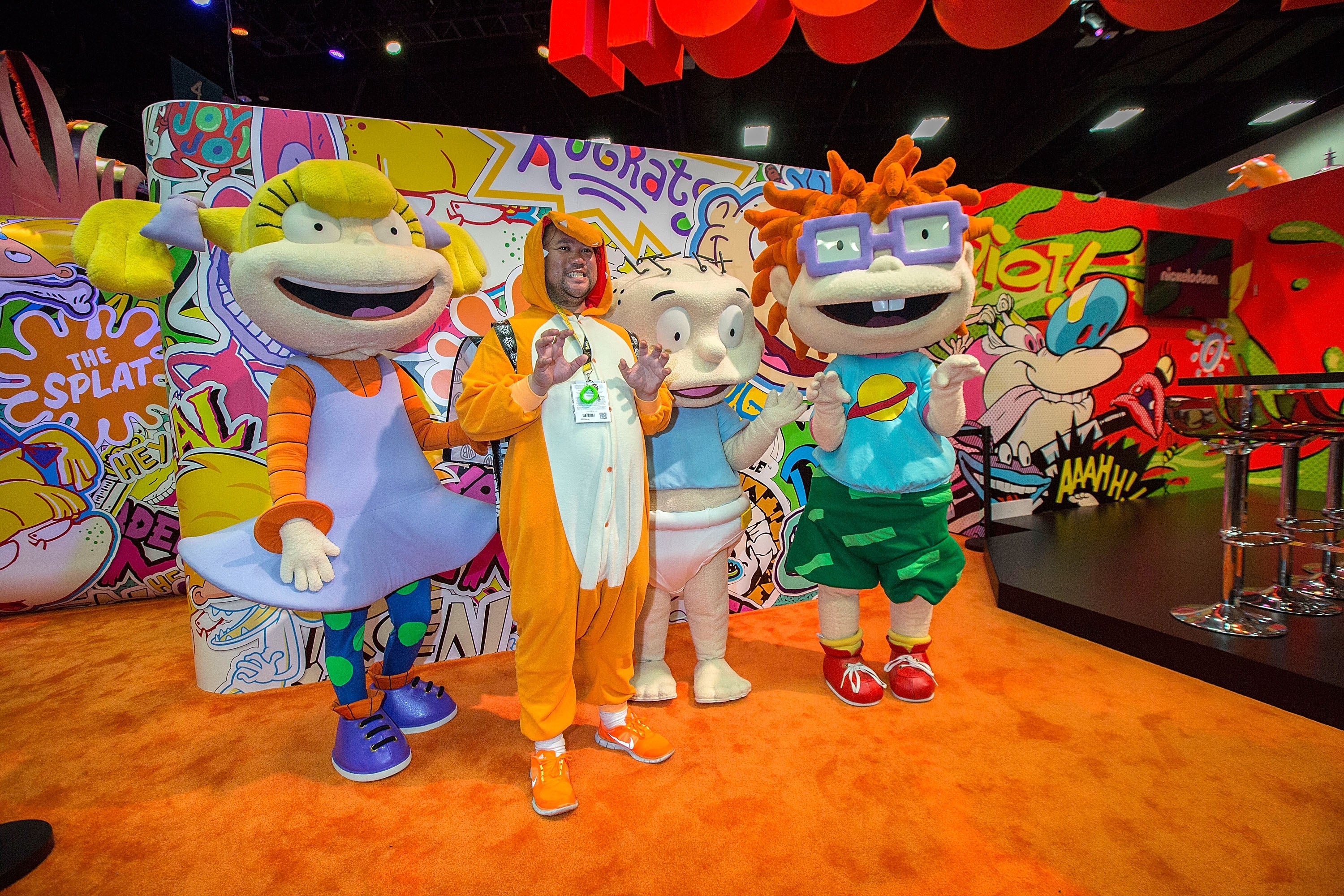 Three radical   dressed arsenic  cartoon characters from 'Rugrats' airs  successful  beforehand   of a colorful backdrop, featuring 90s-themed decor astatine  a convention