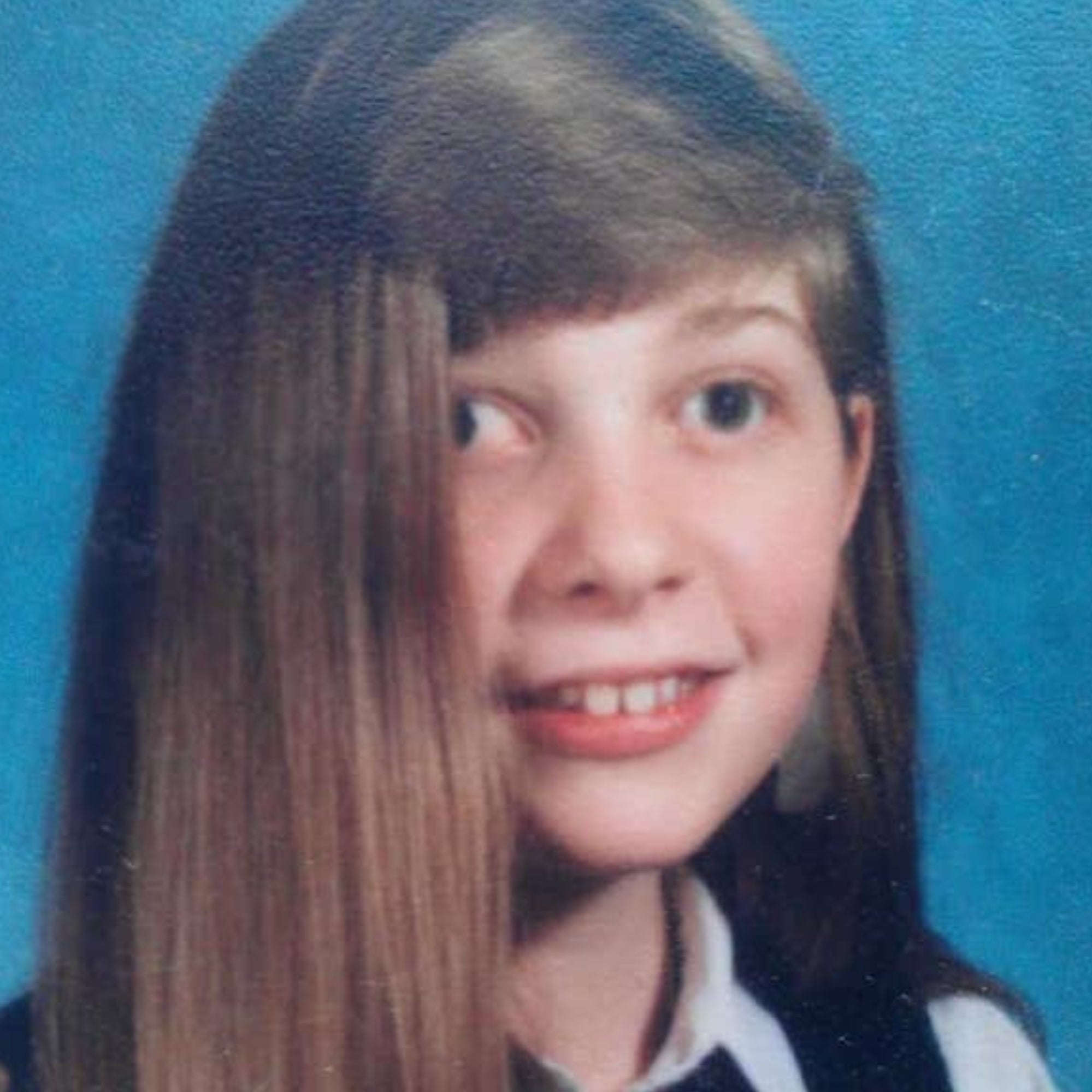 A young person with long straight hair smiles in a school portrait, wearing a collared shirt and a vest
