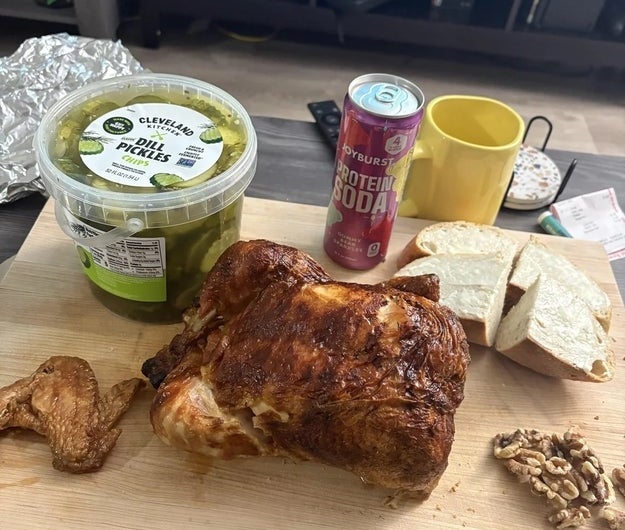 Ayam rotisserie di atas talenan dengan irisan roti, acar, kenari, dan sekaleng soda protein di sampingnya