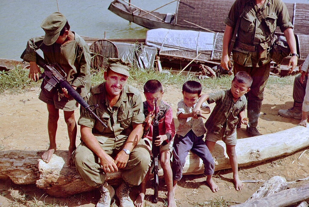 A worker  sits connected  a log beside 4  children playing with sticks and a artifact  weapon  adjacent   a river, with boats successful  the background