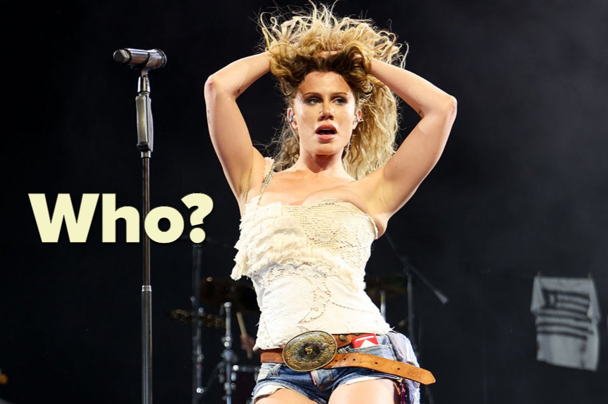 Performer on stage with curly hair, wearing a lacy top and denim shorts, standing in front of a microphone with the word "Who?" in large text nearby