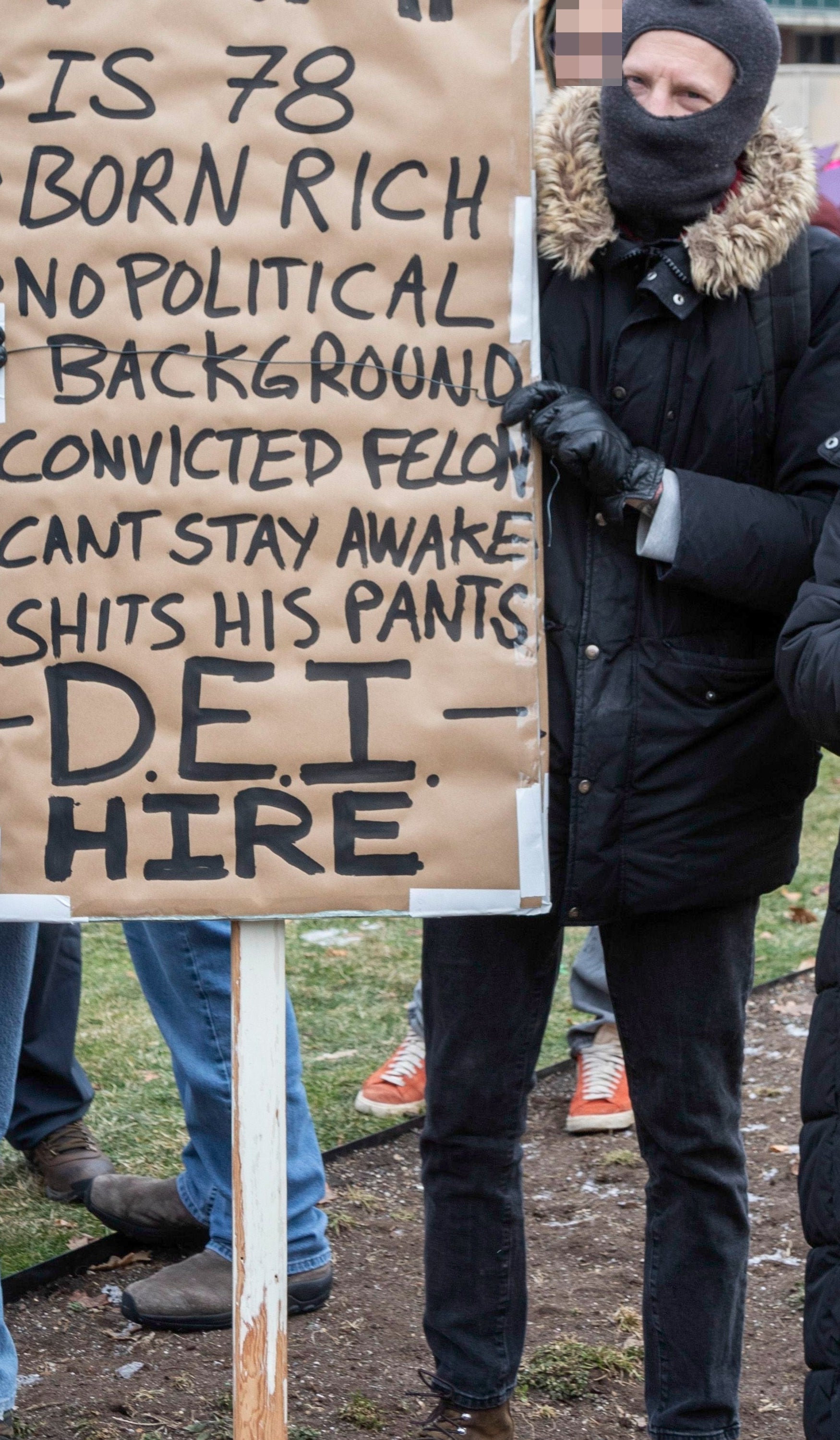 Demonstranter holder et skilt med kritiske uttalelser om en politisk skikkelse, inkludert alder, rikdom og kriminell historie