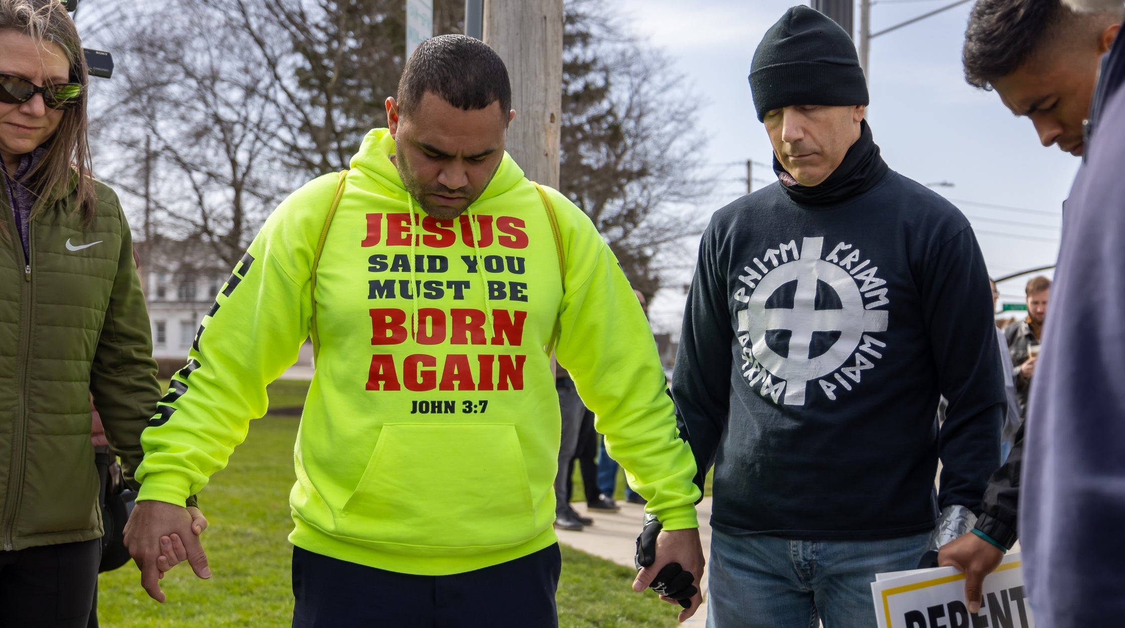 Gruppe mennesker holder hender i bønn utendørs; en person har på seg en genser som leser "Jesus sa at du må bli født på ny, Johannes 3:7."