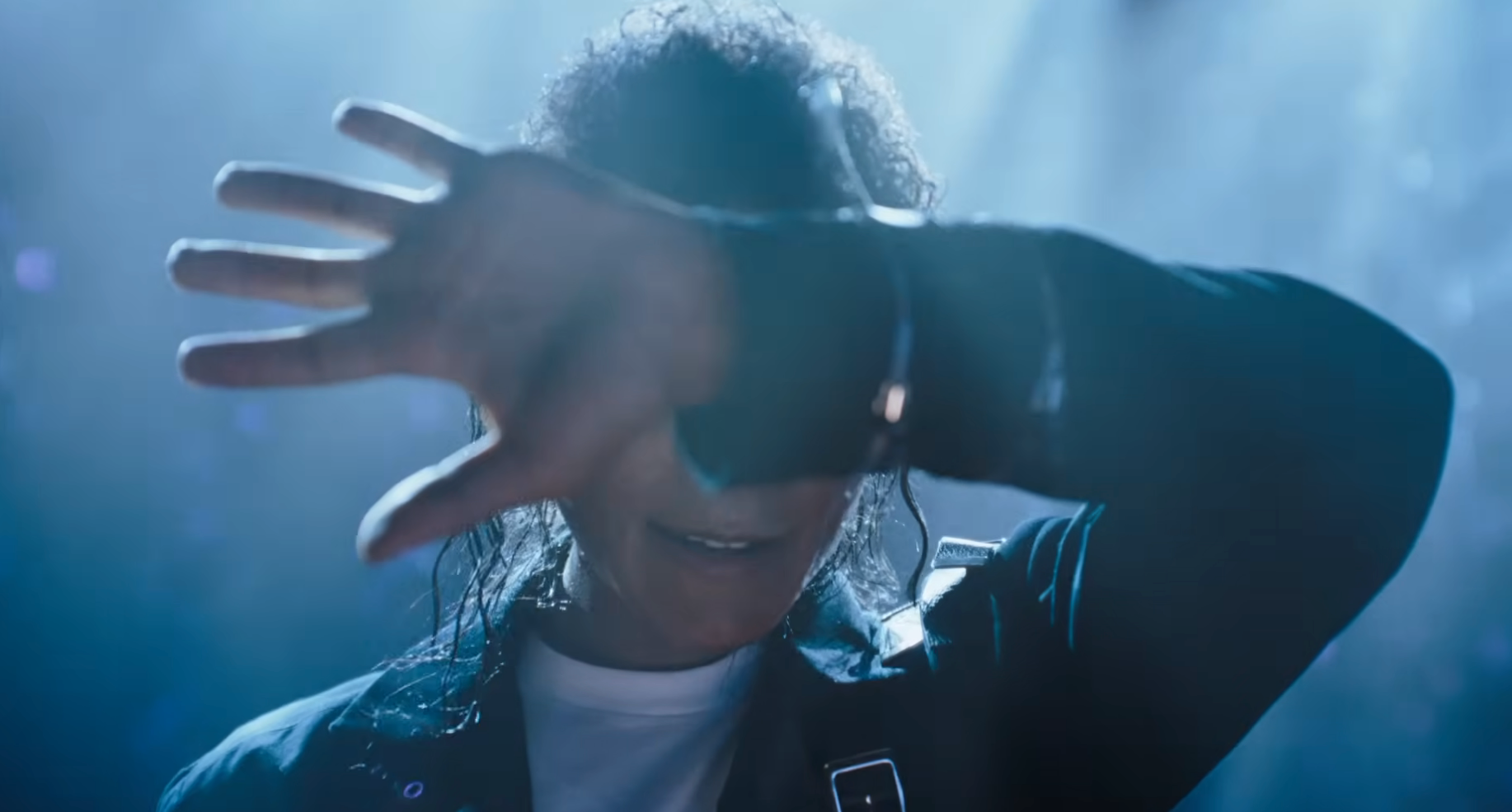 Person connected  stage, partially  obscured by raised arm, wearing a acheronian  overgarment   with buckles. Spotlight creates a melodramatic  effect