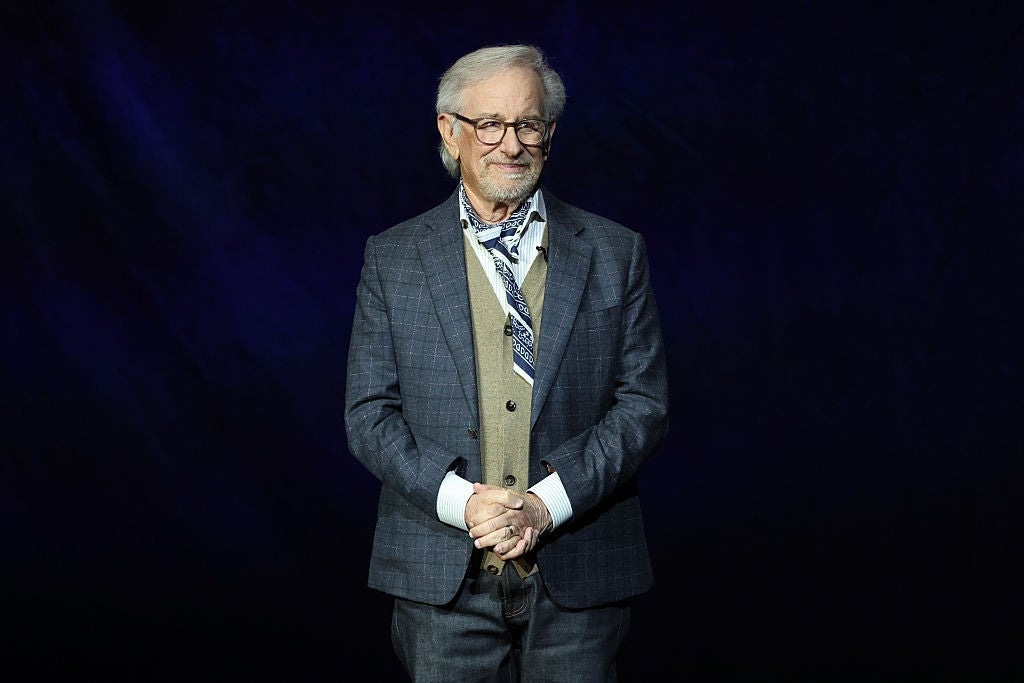 A personage  successful  a checkered suit, vest, and necktie  stands against a acheronian  backdrop, smiling with hands clasped