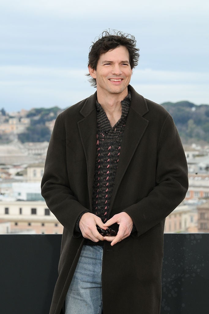 Person smiling outdoors, wearing a agelong  acheronian  overgarment  implicit    a patterned sweater and jeans, with a cityscape successful  the background