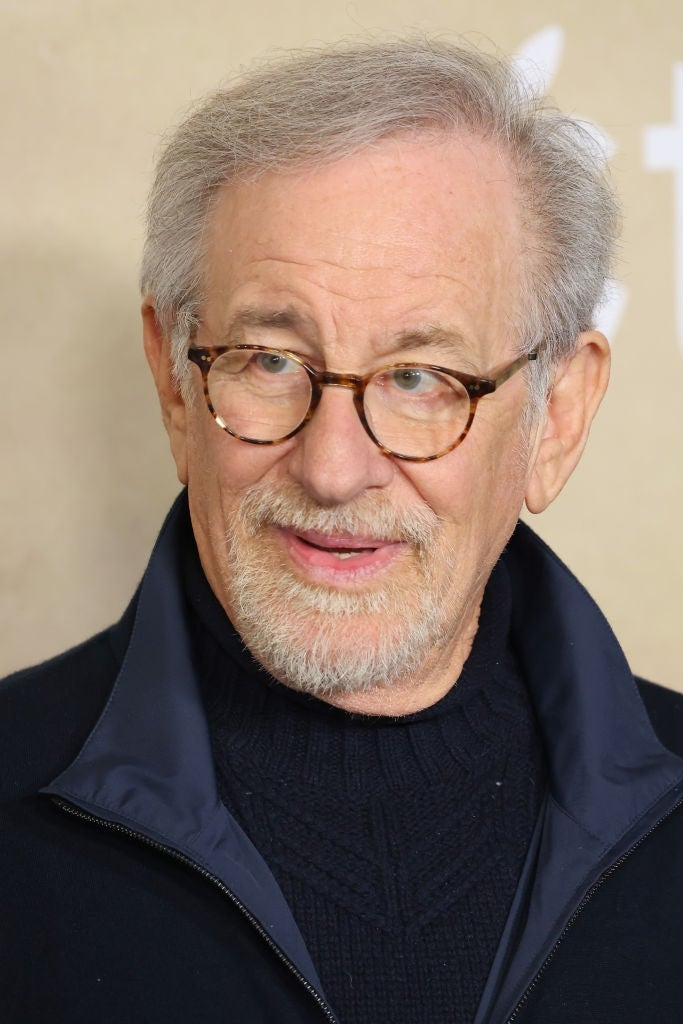 An older man  with glasses, wearing a acheronian  overgarment   implicit    a knit sweater, poses astatine  a personage  event