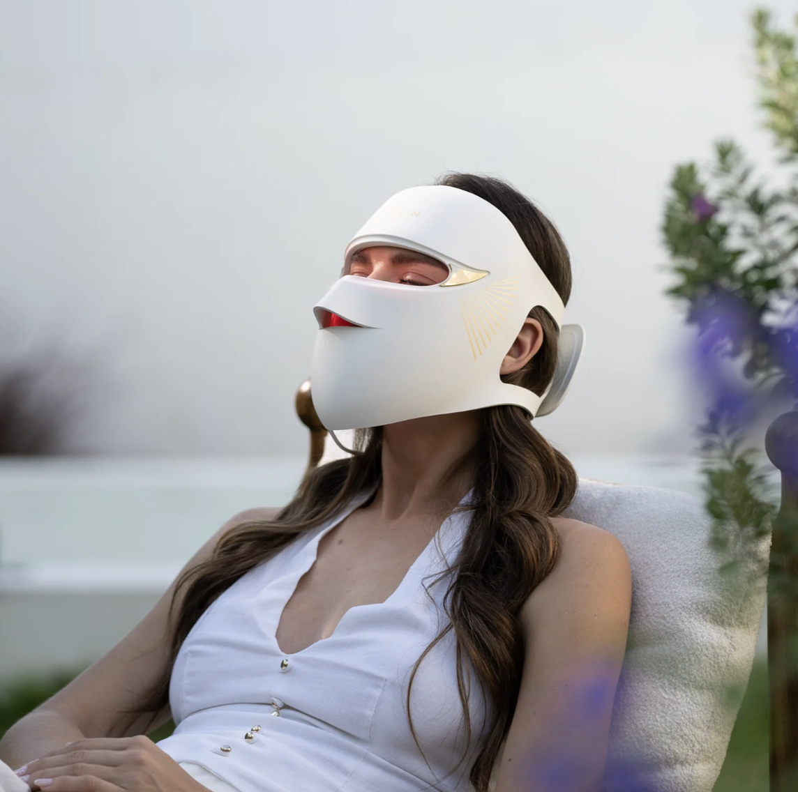 Person relaxing outdoors, wearing a futuristic LED light therapy mask for skincare, dressed in sleeveless white attire