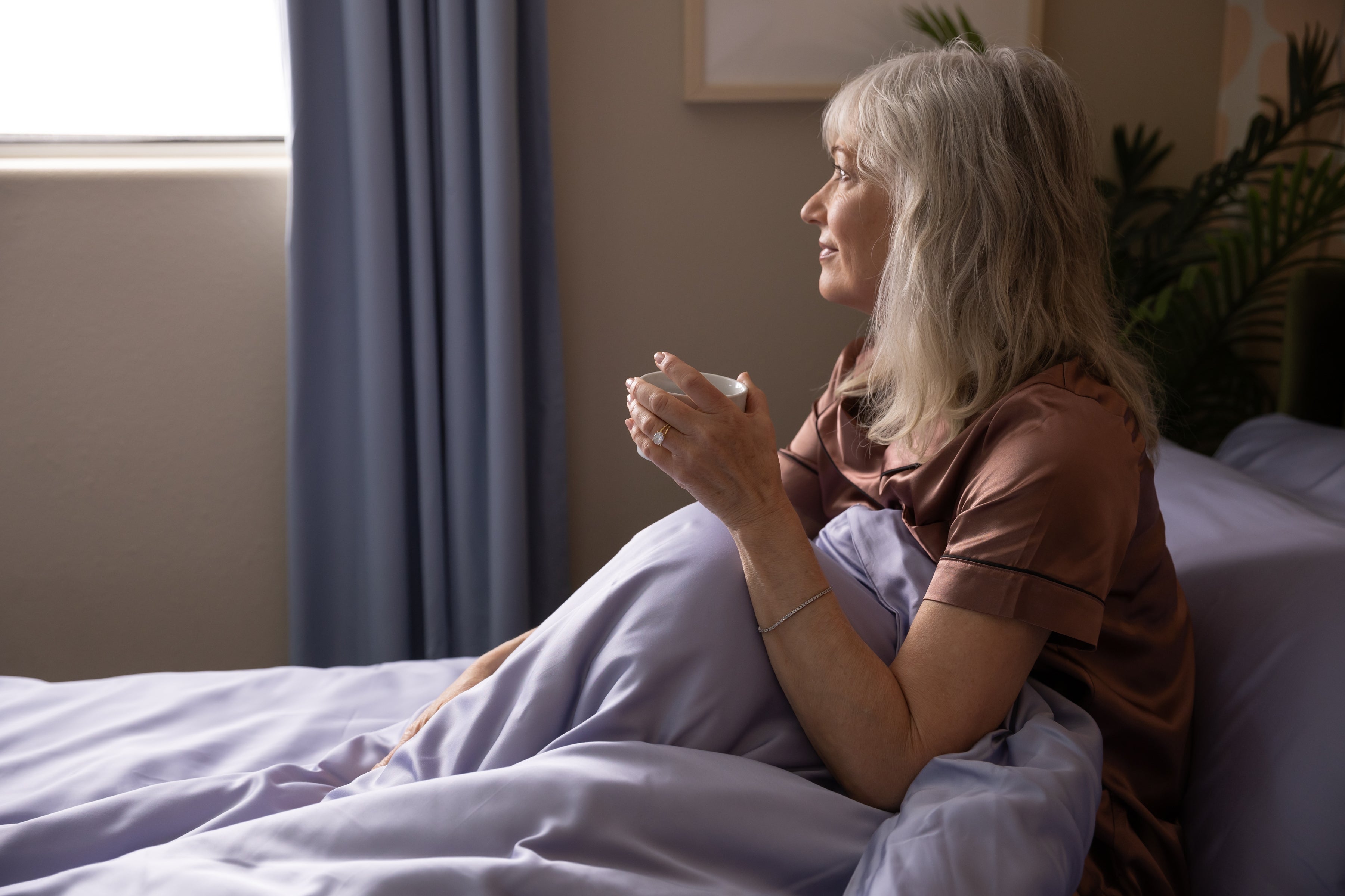 A woman in pajama-style attire sits on a bed, holding a mug and looking out a window, suggesting a calm morning routine