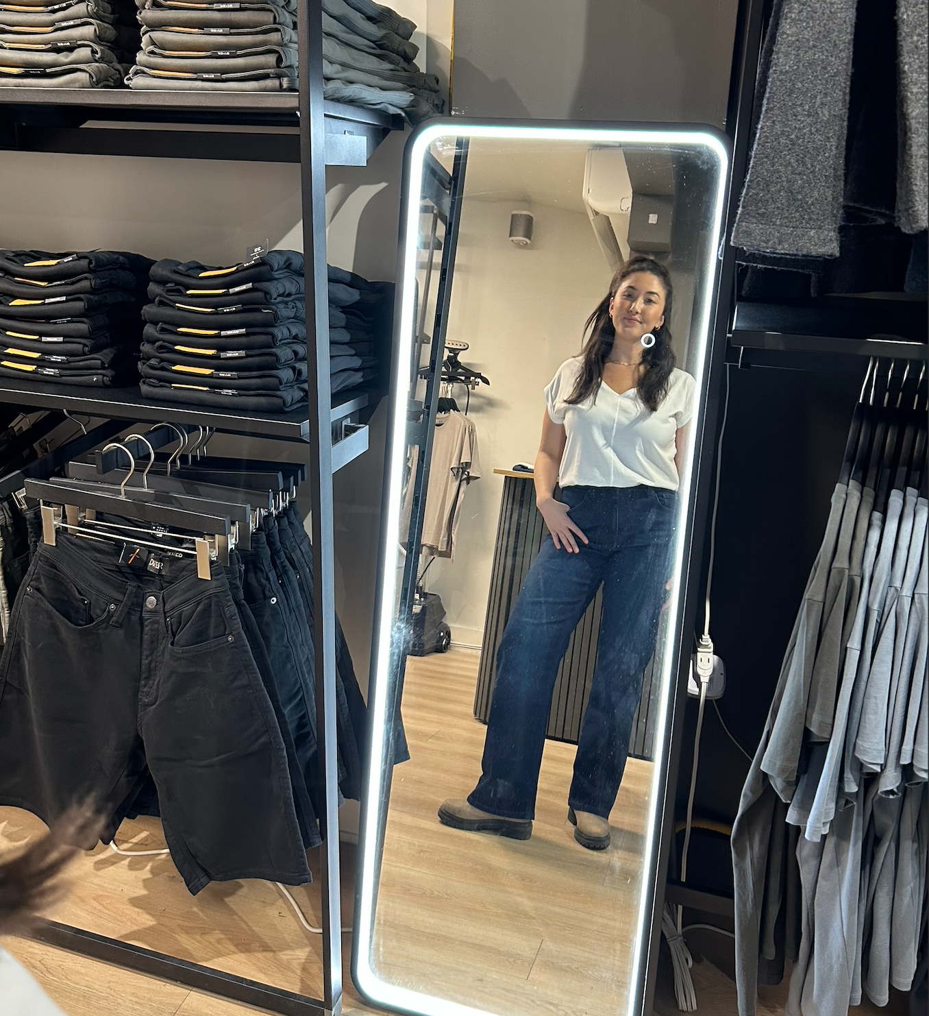 Woman in a stylish casual outfit—white top, blue jeans, brown shoes—stands in a clothing store, reflected in a tall mirror, surrounded by folded clothes