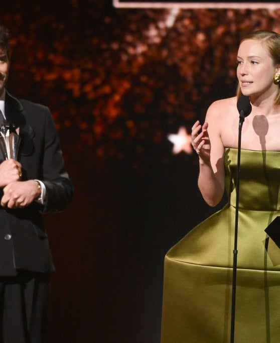 Two radical   connected  signifier    astatine  an grant  show; 1  successful  a achromatic  suit   holding a trophy, the different   successful  a geometric formal  speaking into a microphone